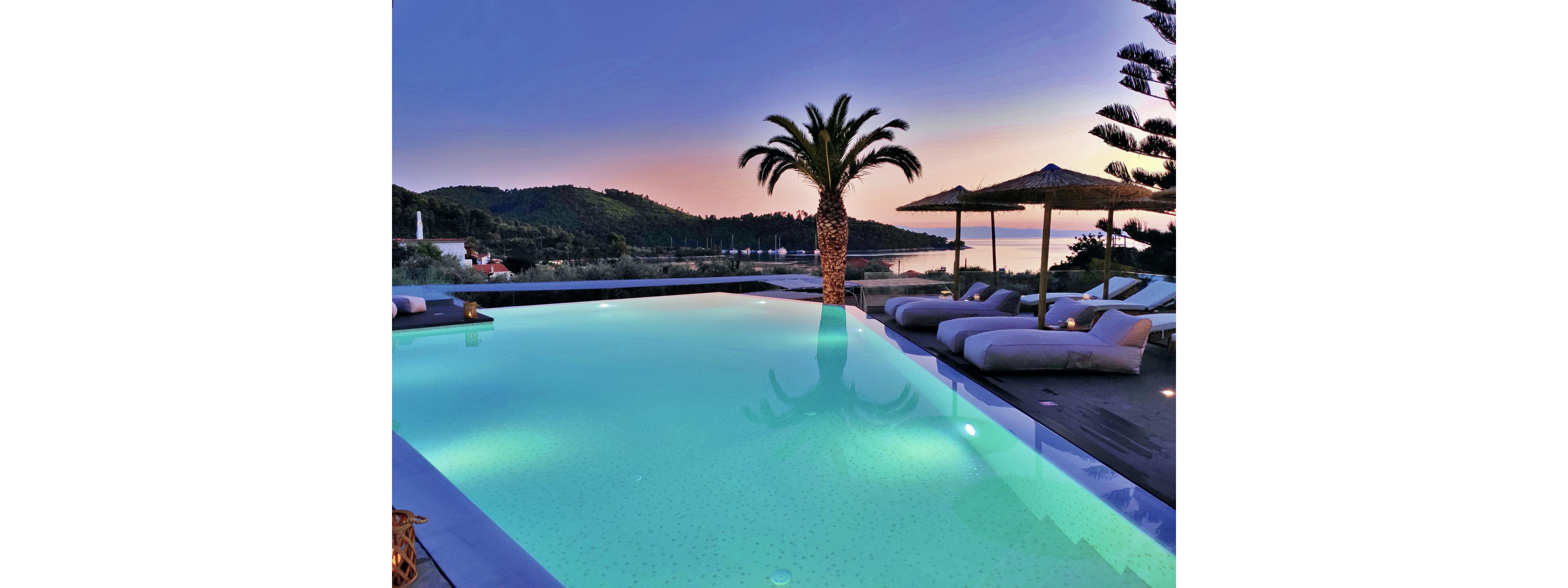 A serene infinity pool glows with turquoise light at dusk, overlooking a calm coastal landscape. Comfortable lounge chairs and straw parasols line the poolside deck, inviting relaxation. A tall palm tree stands prominently against the pastel sky, while distant hills and water create a tranquil backdrop. The scene captures a luxurious resort atmosphere with a peaceful, tropical ambiance.