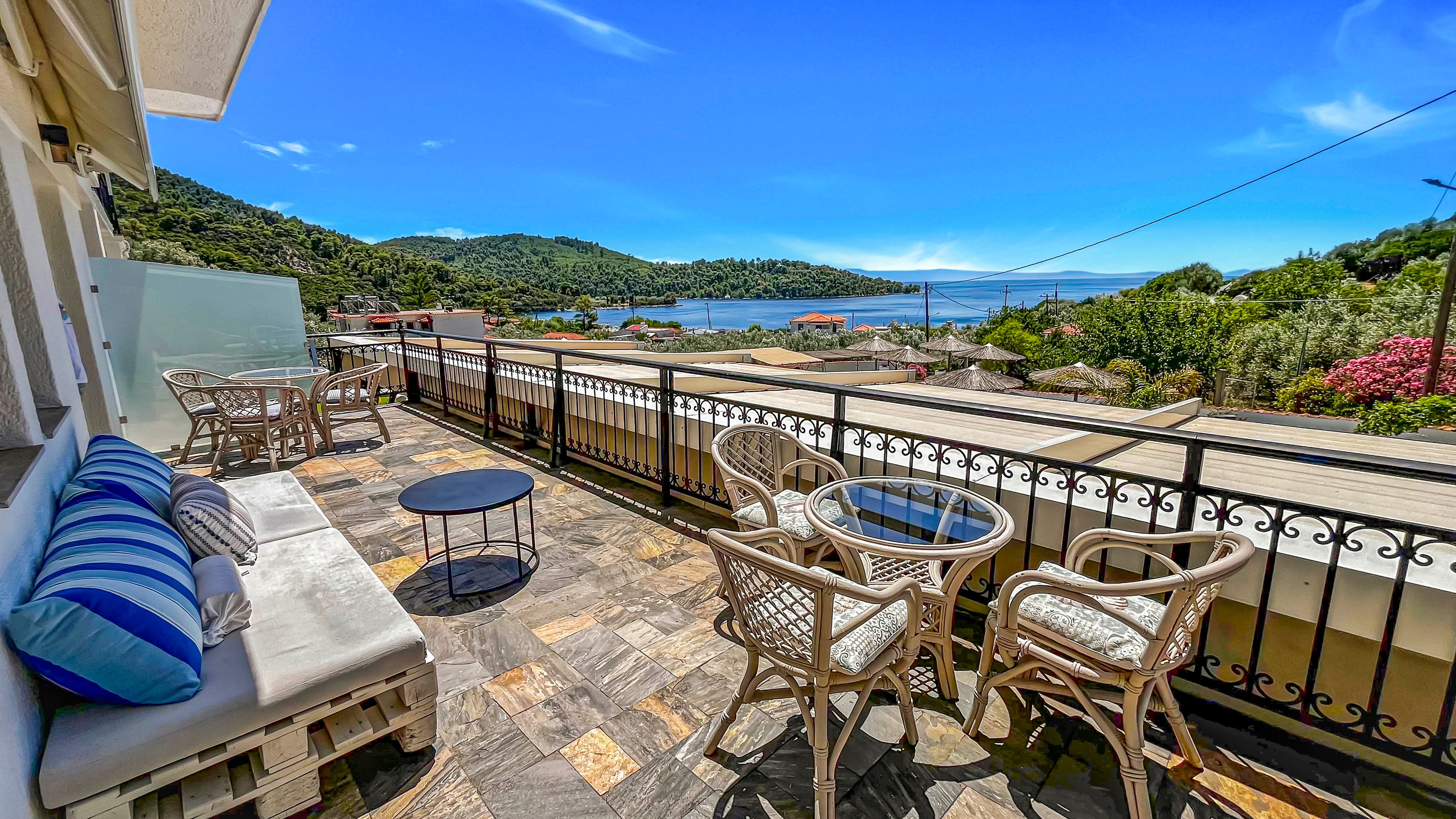 A spacious terrace overlooks a coastal village and blue sea under a clear sky. Wicker chairs and glass-topped tables are arranged beside a cushioned bench with striped pillows. The tiled floor and black metal railing frame the scenic view of green hills and scattered rooftops. Bright daylight and vivid colors create a relaxed, vacation-like atmosphere.