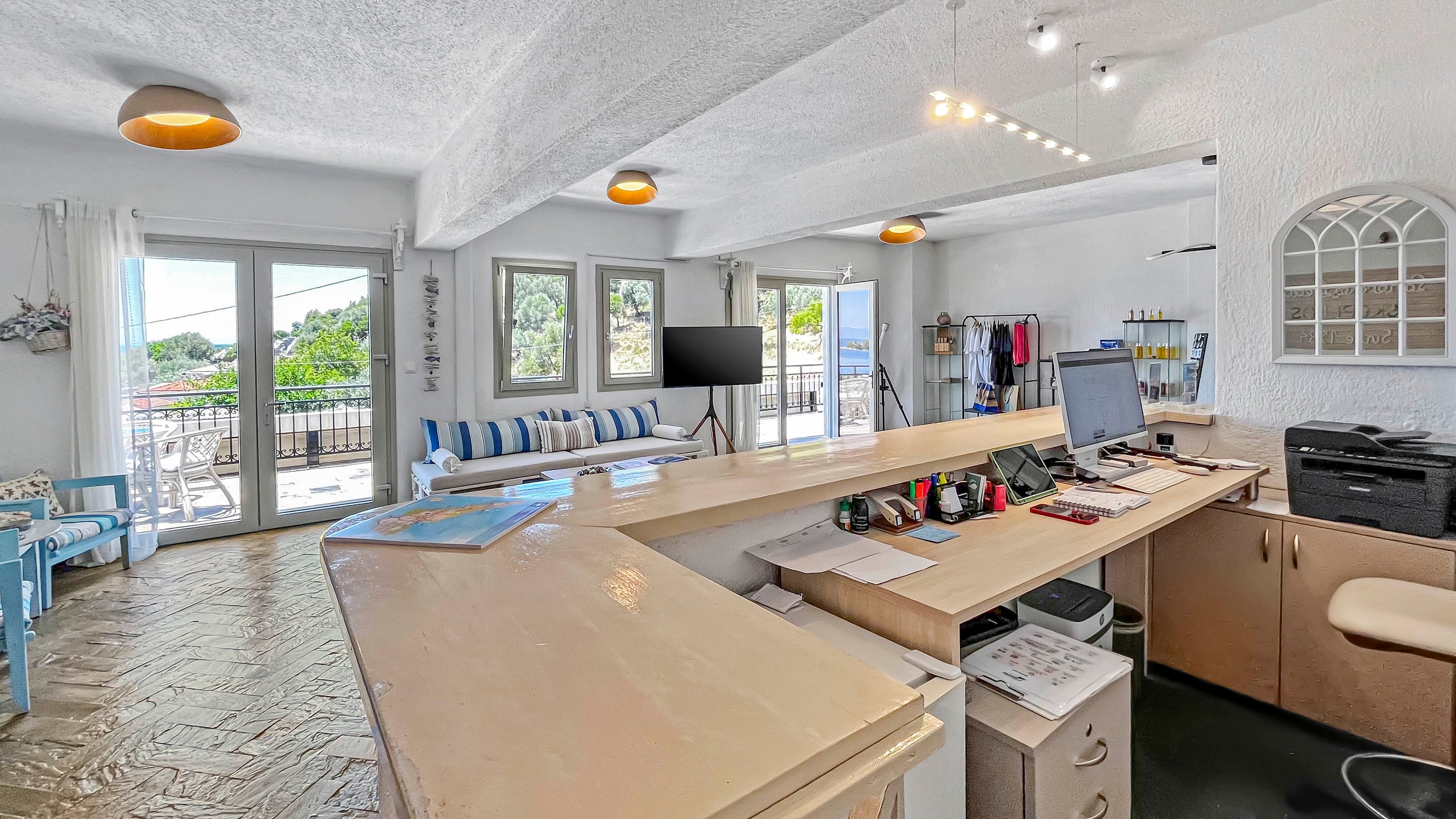 A bright office reception area features a long front desk with a computer, printer, and office supplies neatly arranged. Behind the desk, a cozy seating area with striped cushions faces several windows and glass doors that open to a balcony with greenery outside. The white walls, light wood surfaces, and tiled floor create an airy, coastal atmosphere. Track lighting and ceiling fixtures illuminate the space, highlighting both the work area and the lounge seating.