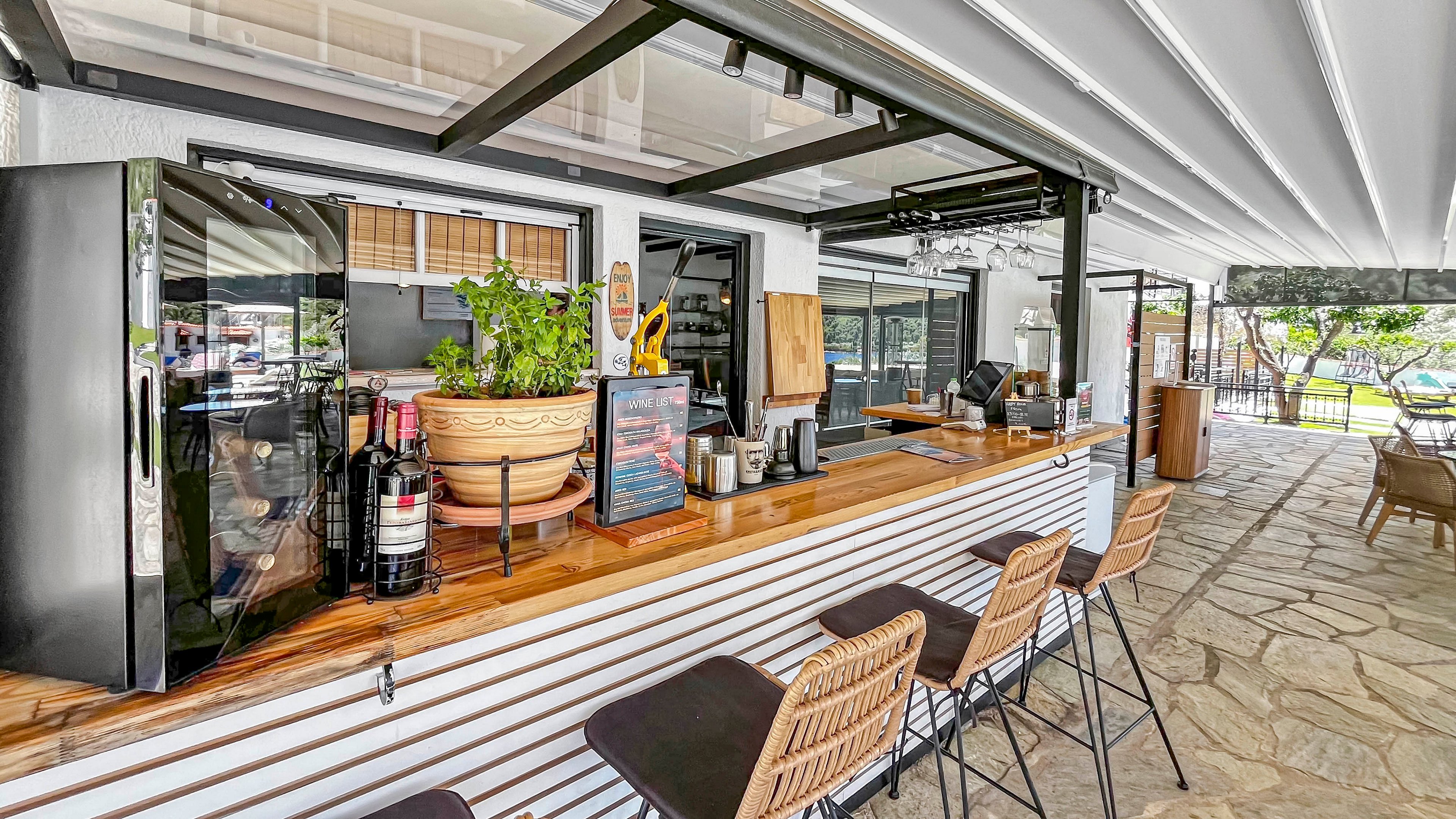 An outdoor cafe bar counter features a wooden surface with several bottles, plants, and drink-making equipment neatly arranged. High stools line the front of the bar, inviting customers to sit and enjoy refreshments. The bar is sheltered by a modern covered patio structure, with a stone floor extending into the seating area. In the background, additional tables and chairs overlook a bright, green outdoor space.