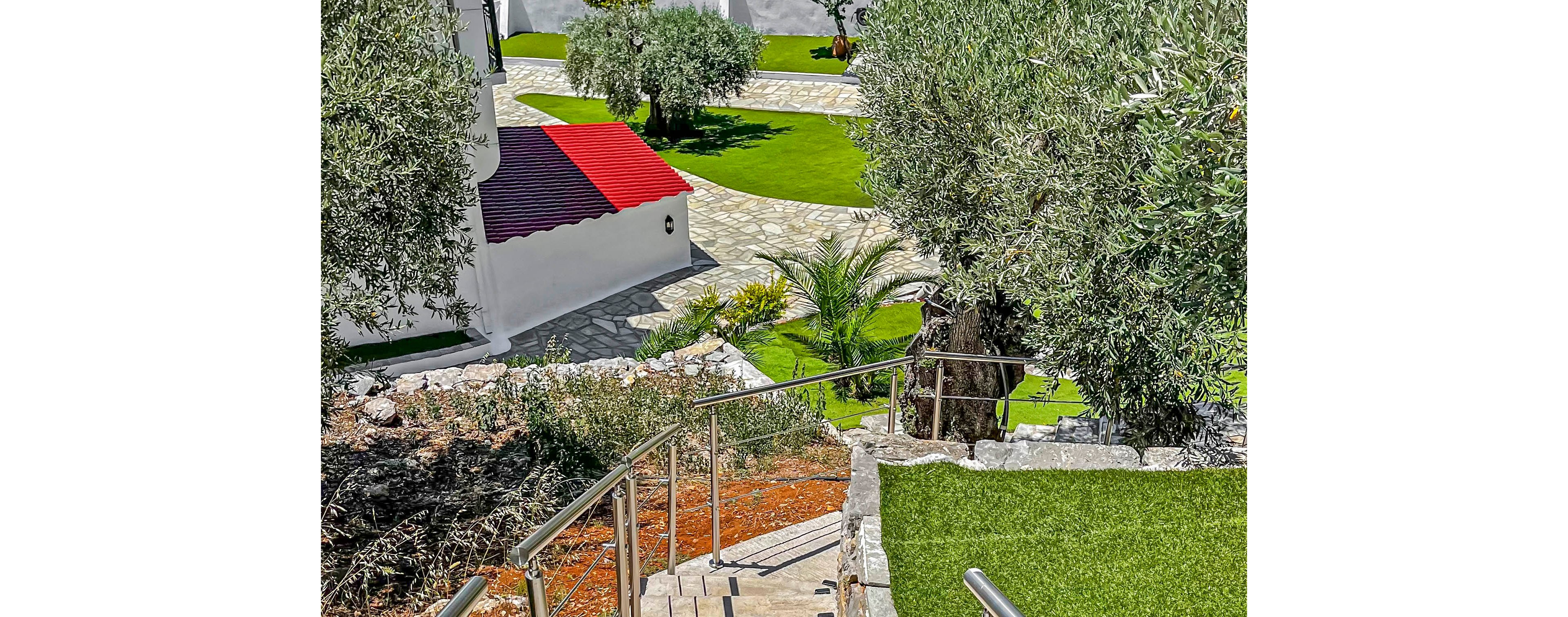The image shows a stone stairway leading down to a landscaped garden area beside a white house with a red roof. Curved stone paths wind through bright green lawns and small ornamental plants. Metal railings line the stairs, and mature trees provide shade around the property. The overall scene suggests a peaceful residential or vacation setting with well-maintained outdoor spaces.