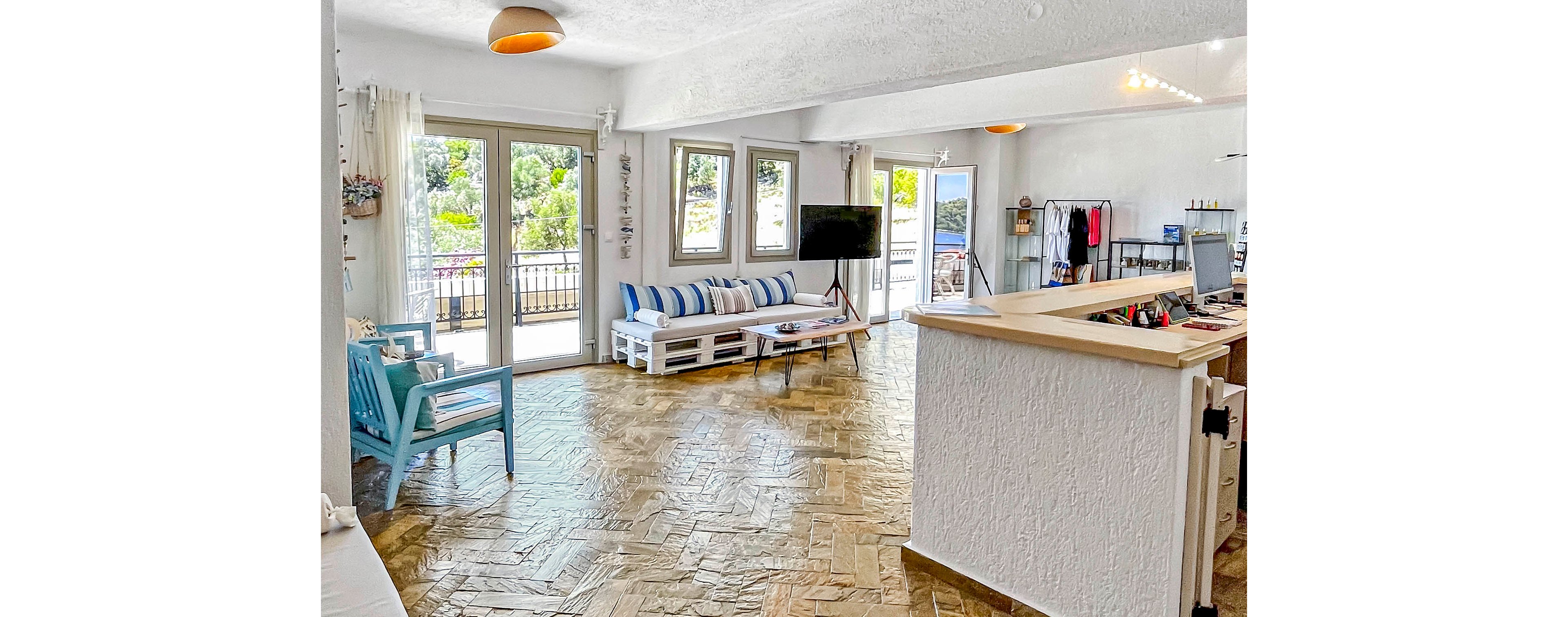 Spacious open-plan living room with light wood flooring and white textured walls. Large glass doors and multiple windows open to a balcony overlooking greenery and trees. A white sofa with blue striped cushions and a turquoise armchair create a coastal seating area facing a flat-screen TV. The room includes a small bar or counter space, minimal decor, and abundant natural light for an airy, relaxed atmosphere.