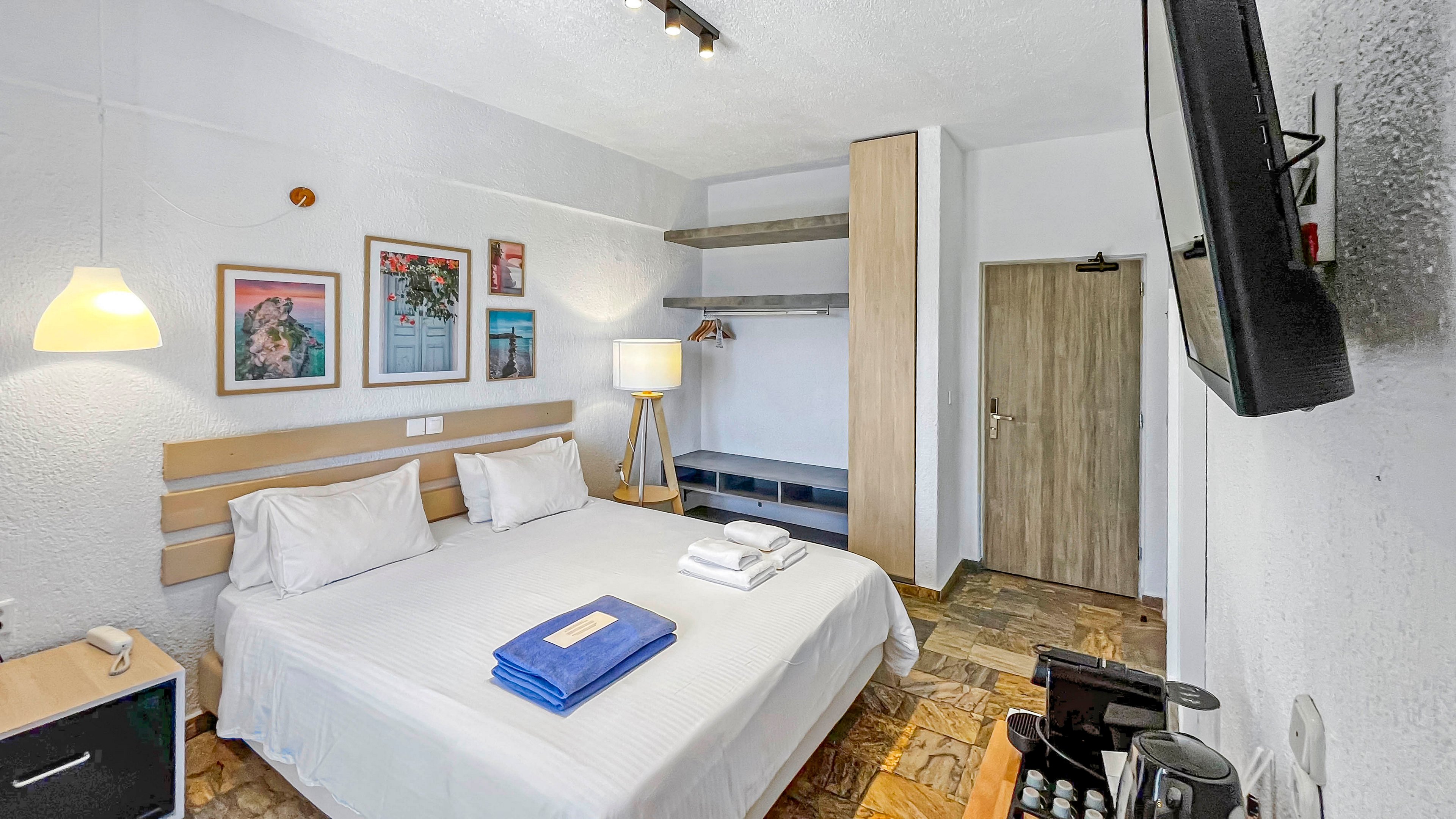 A clean, modern hotel room features a neatly made double bed with white linens and folded towels and a blue blanket placed on top. The space includes wall art above the headboard, a wooden wardrobe with open shelving, and a floor lamp providing warm light. A flat-screen television is mounted on the wall opposite the bed, and a small refreshment station is visible in the foreground. The room has a bright, inviting atmosphere with neutral walls and a patterned floor.