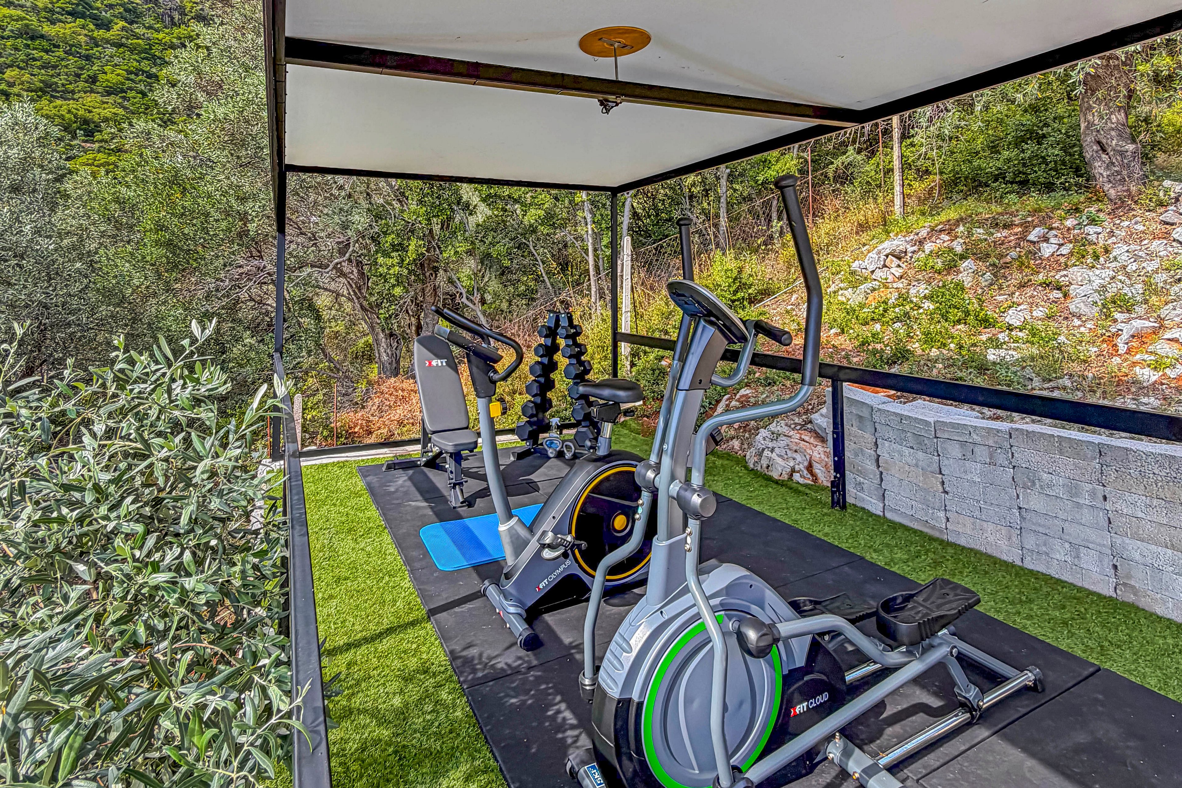 An outdoor gym setup features an elliptical trainer and an exercise bike under a covered canopy. A weight bench and a vertical dumbbell rack stand nearby, providing strength training options. The space is finished with artificial grass flooring and bordered by a low stone wall. Lush green trees and hillside vegetation surround the fitness area, creating a natural, scenic workout environment.