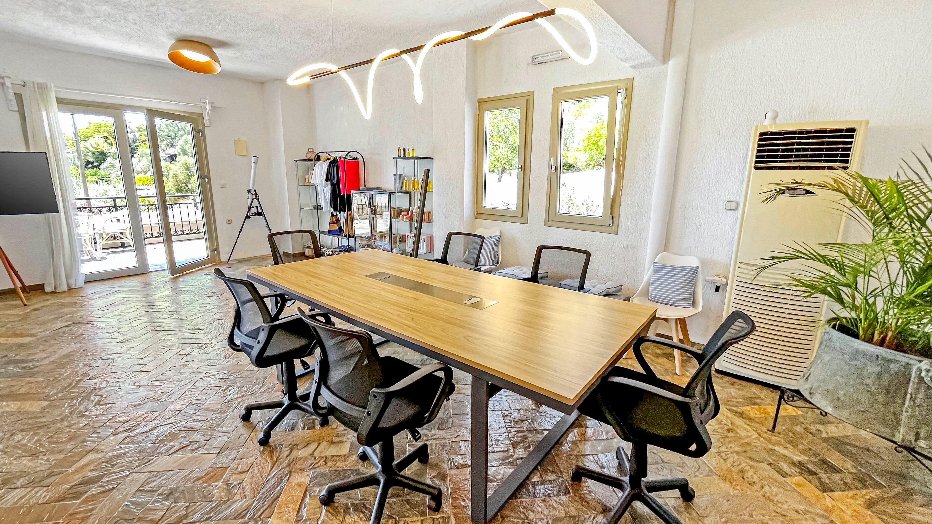 A contemporary meeting room features a long wooden conference table surrounded by black office chairs. Large windows and glass doors let in abundant natural light and offer a view of greenery outside. The space includes a freestanding clothes rack, a tripod, and a wall-mounted air conditioner, adding functional elements to the room. Warm lighting fixtures and indoor plants contribute to a relaxed, creative workspace atmosphere.