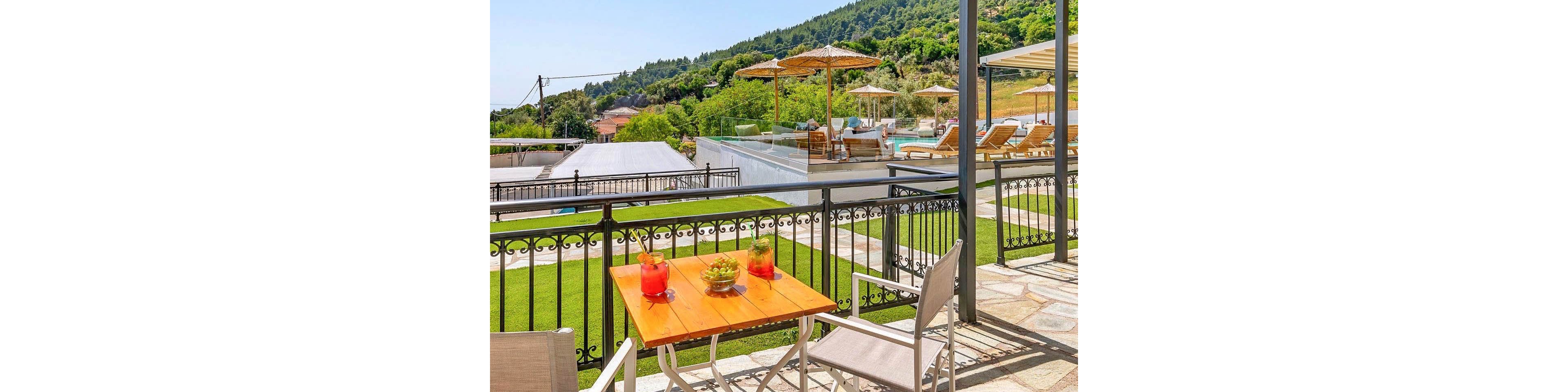 A bright terrace with a small wooden table and chairs overlooks a resort pool area and lush green hills. Two colorful drinks and a small potted plant sit on the table, suggesting a relaxing outdoor refreshment. Sun loungers and umbrellas surround the pool in the background, framed by decorative metal railings. The scene is bathed in warm daylight, creating a tranquil vacation atmosphere.