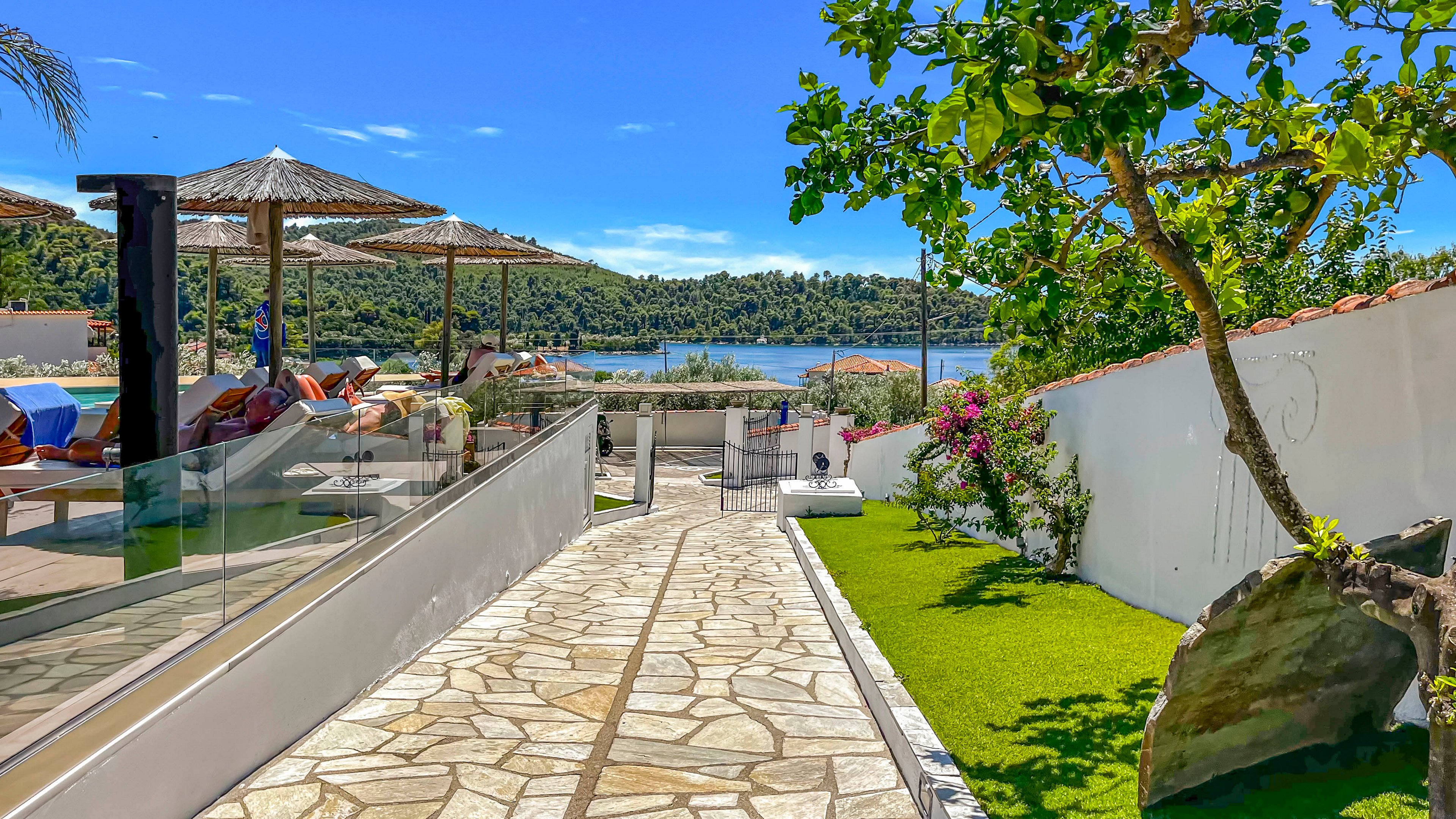 A stone pathway leads through a landscaped garden toward a bright lakeside terrace. Sun loungers and straw parasols line the left side, separated by a glass railing. On the right, a manicured green lawn, flowering shrubs, and a small tree add vibrant color. In the background, calm blue water and forested hills create a tranquil vacation setting under a clear sky.