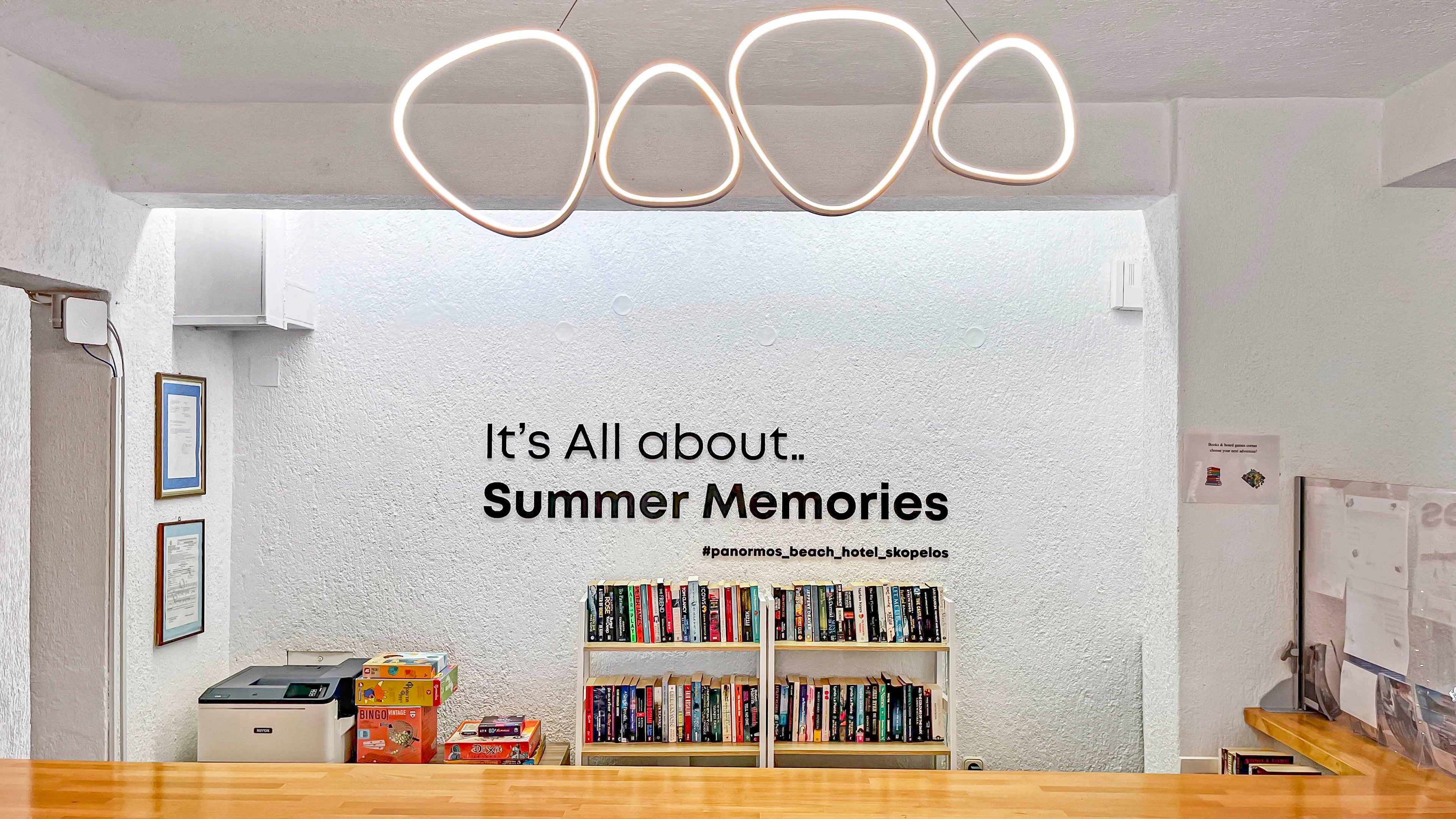 A bright hotel lobby features a wooden counter in the foreground and a white wall with a bookshelf filled with colorful books. Above the shelf, the text reads "It's All about.. Summer Memories" along with a social media handle. Modern loop-shaped ceiling lights hang from the white ceiling, adding a contemporary touch. A printer and some board games sit on a side table, suggesting a relaxed, welcoming atmosphere for guests.