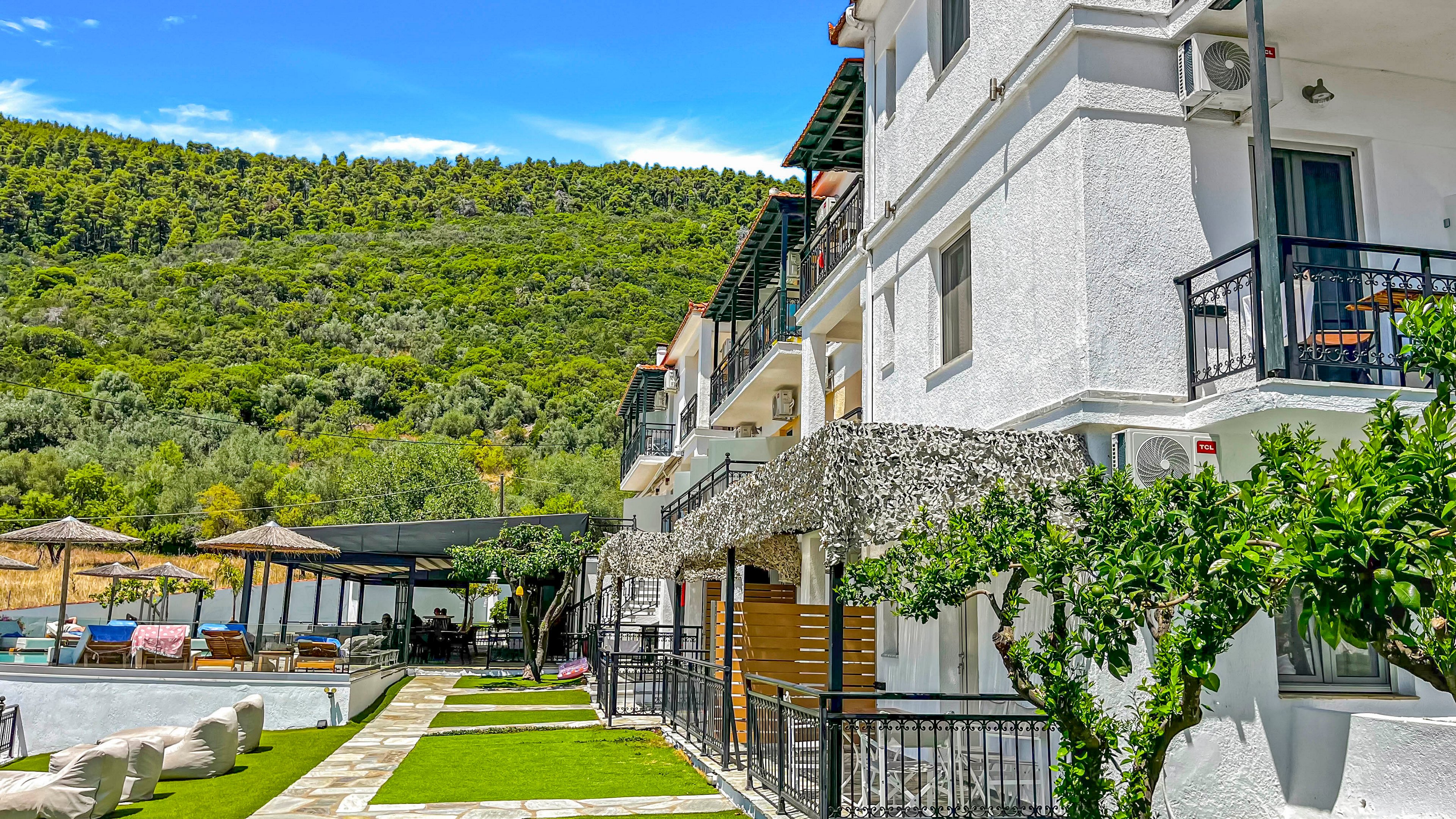 A white multi-story hotel building stands beside a landscaped pool area under a bright blue sky. Green hills covered with trees rise in the background, creating a scenic natural setting. Sun loungers, umbrellas, and shaded seating areas line the poolside, suggesting a relaxing vacation atmosphere. Balconies, potted plants, and trimmed grass add to the inviting resort feel.