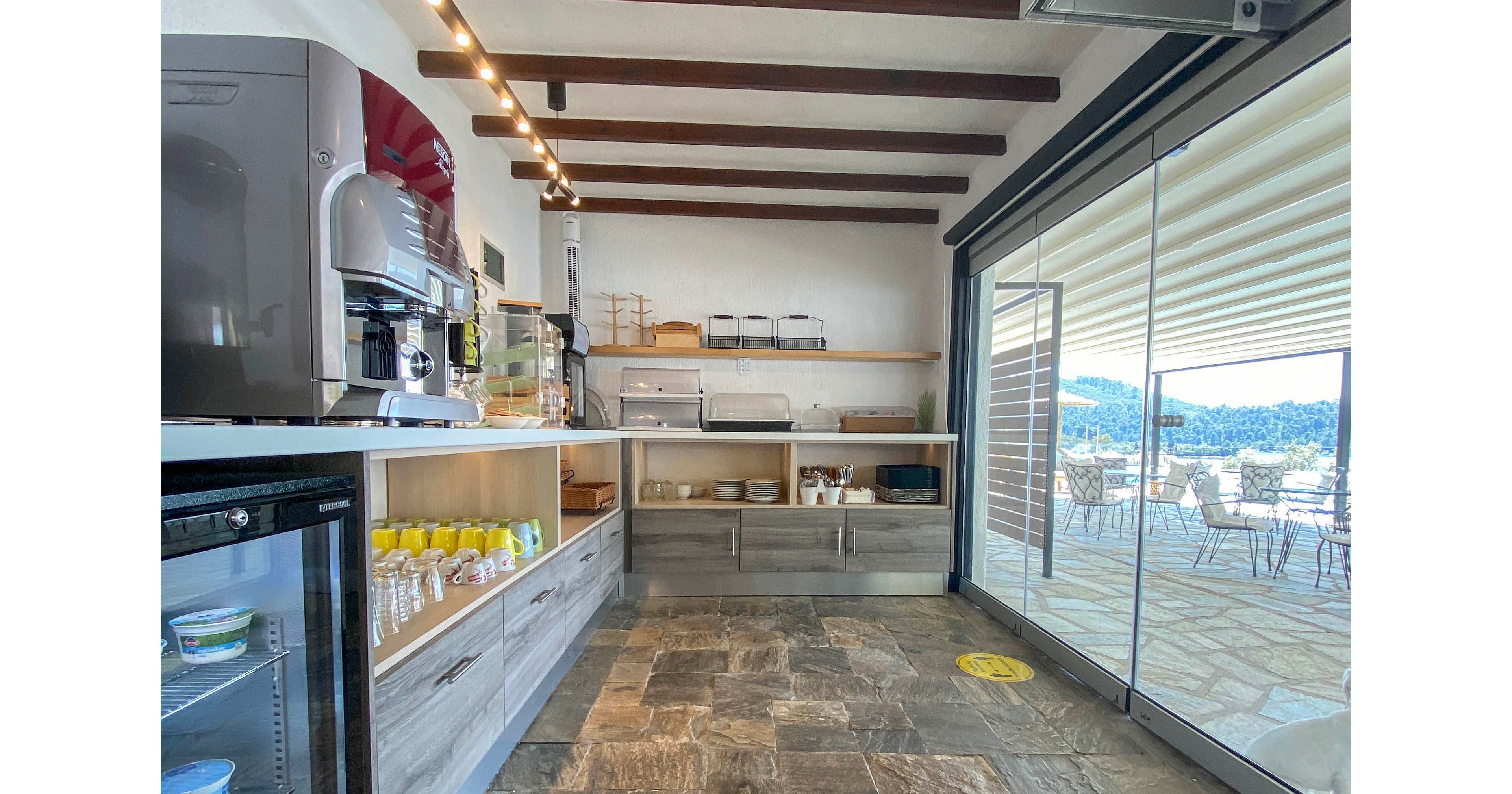 Bright self-service cafe area with stone tile flooring and wooden ceiling beams. Countertops display cups, dishes, and food service equipment, including a coffee machine and refrigerated unit. Large glass sliding doors open to an outdoor seating terrace overlooking a scenic hillside landscape. The space appears clean, organized, and contemporary, with natural light enhancing the airy atmosphere.
