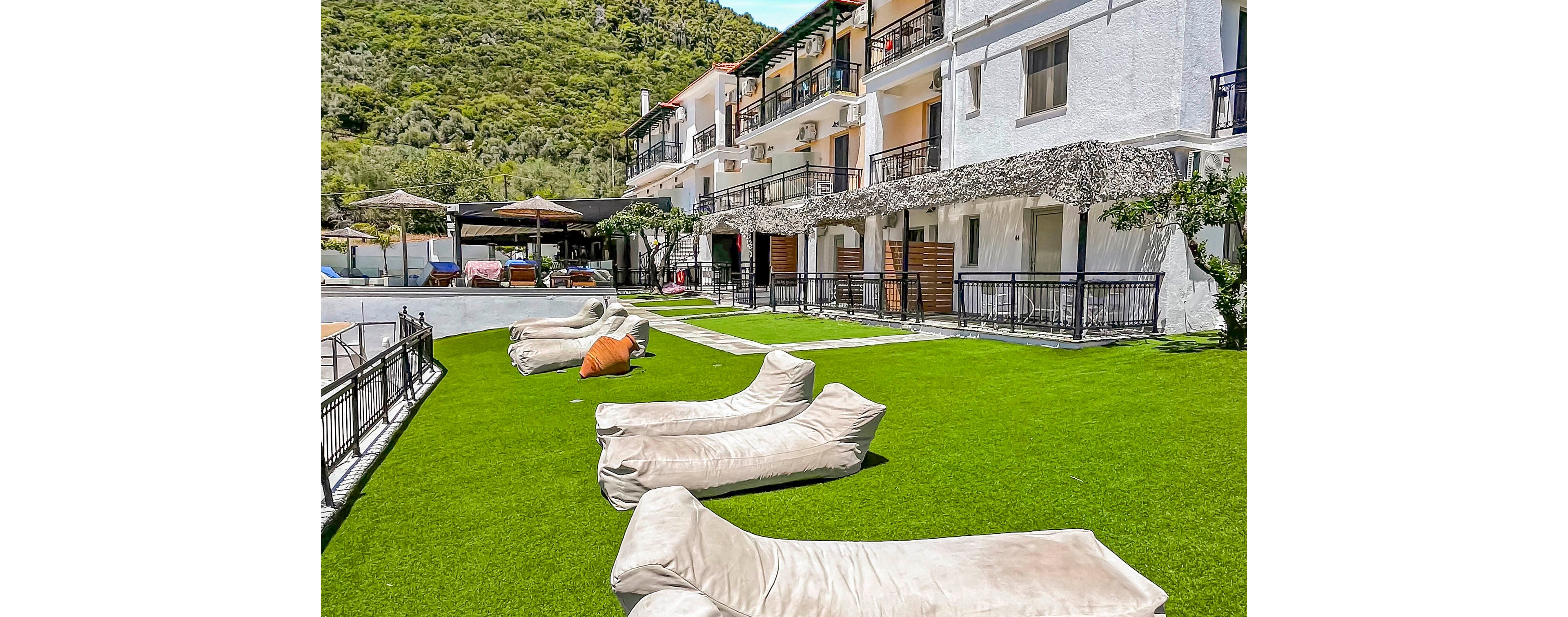 A bright resort courtyard features several beige lounge chairs arranged on a vivid green lawn. White multi-story buildings with balconies line one side, facing the open relaxation area. In the background, sun umbrellas, outdoor seating, and a poolside zone suggest a vacation setting. Lush green hills rise behind the property, adding a natural backdrop to the sunny, inviting atmosphere.