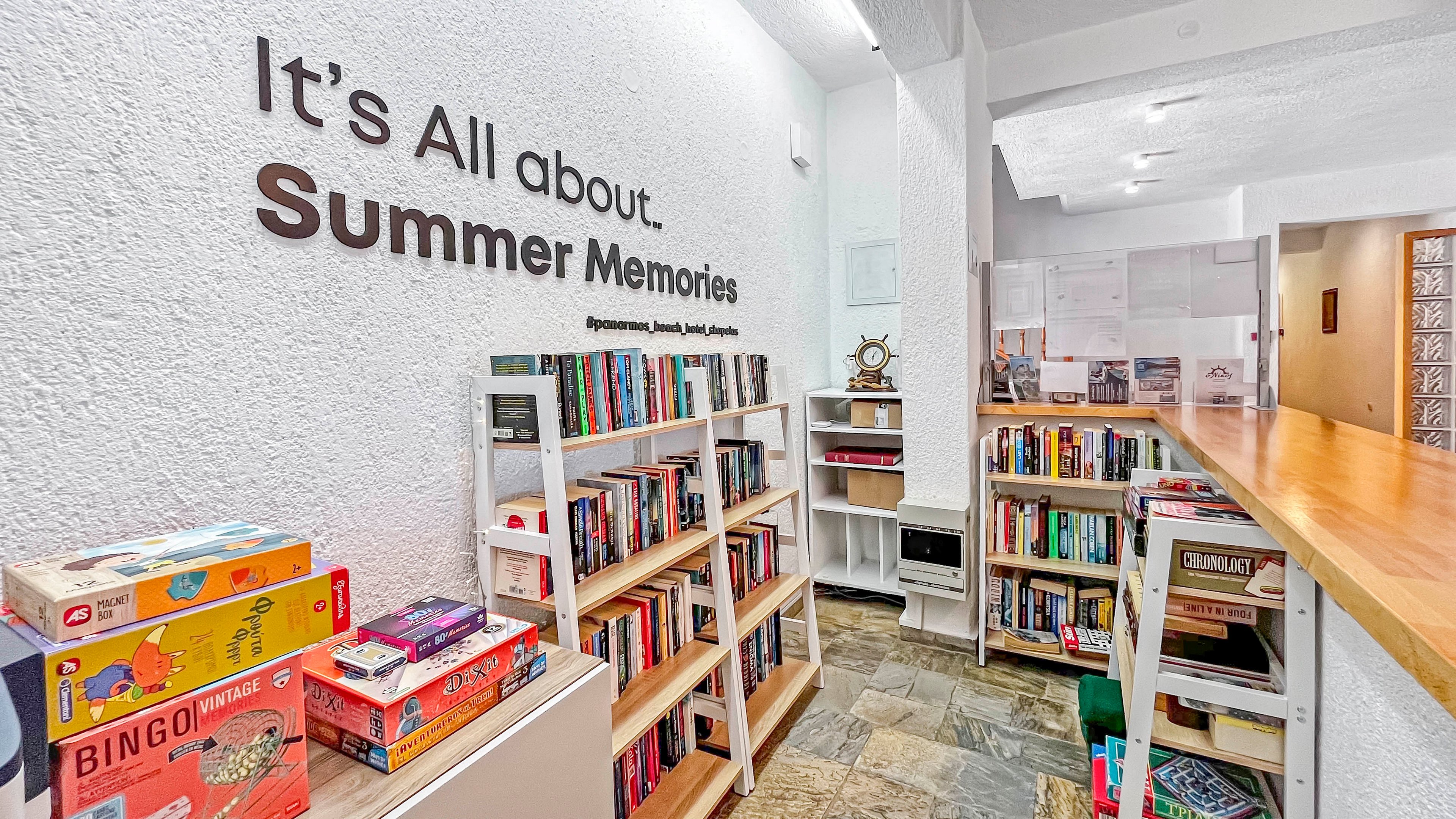 A bright indoor reading corner features white bookshelves filled with assorted books and board games. On the textured wall, large text reads "It's All about Summer Memories" above the shelves. Colorful game boxes, including one labeled "BINGO", add playful accents to the scene. The space has a warm, inviting atmosphere with a long wooden counter and stone-tile flooring extending into the background.