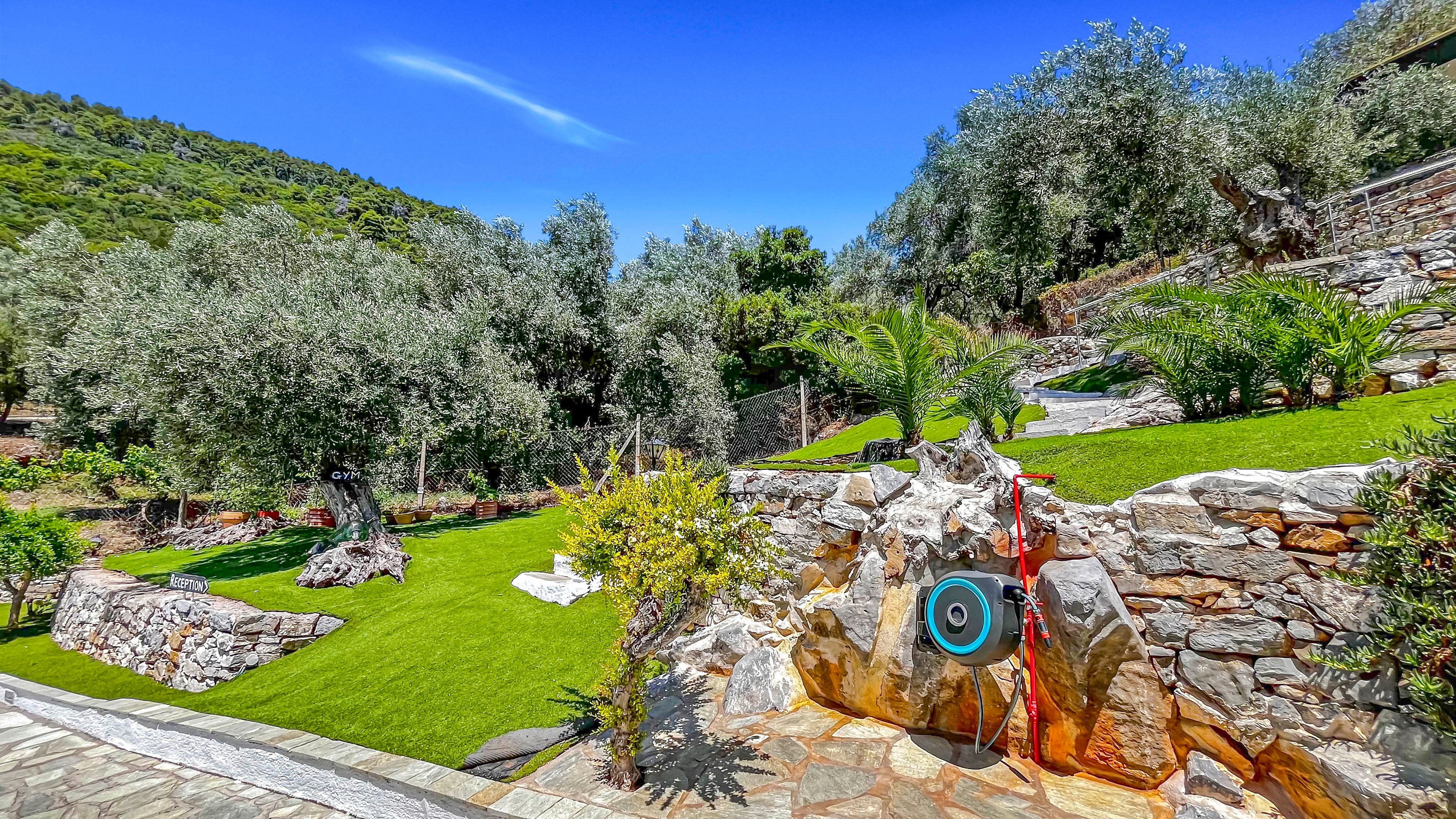 A terraced garden is set on a hillside with neatly maintained green lawns and stone retaining walls. Mature trees and shrubs surround the area, creating a lush Mediterranean landscape under a clear blue sky. A circular target or decorative element is mounted on a rock near a red hose, adding a focal point in the foreground. The scene feels bright and tranquil, emphasizing the contrast between natural stone, vibrant grass, and dense foliage.