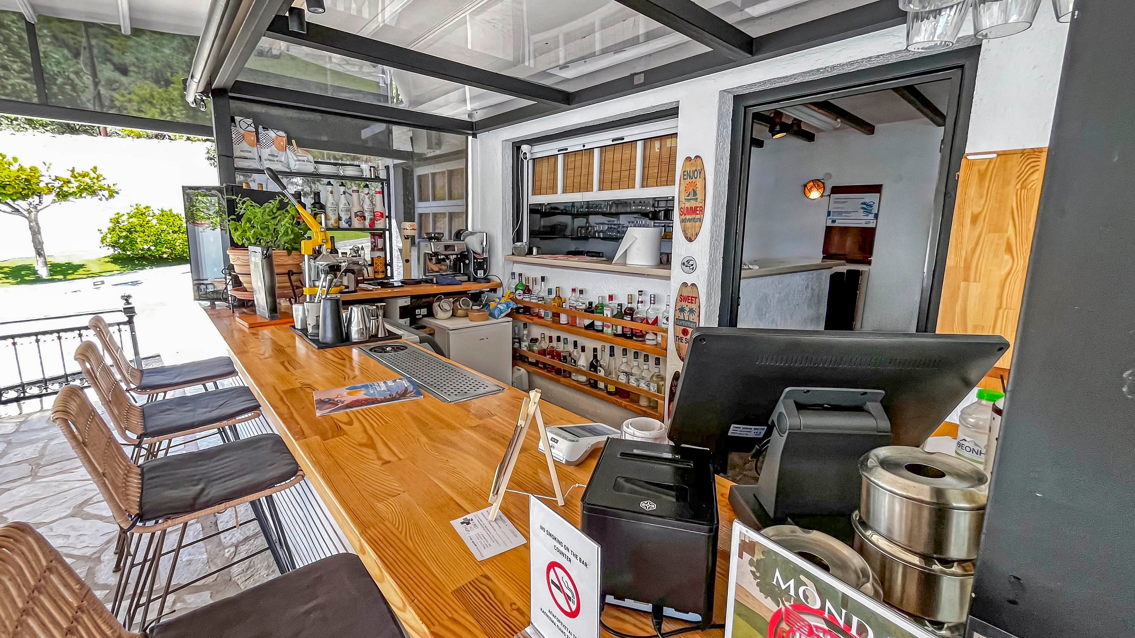 A modern open-air bar features a long polished wooden counter with several stools neatly arranged along it. The back wall displays shelves filled with various bottles and glassware, while a small service window opens into an interior room. A point-of-sale terminal and metal containers sit on the counter near a sign with a red prohibition symbol. Bright natural light and greenery outside suggest a relaxed coastal or resort setting.