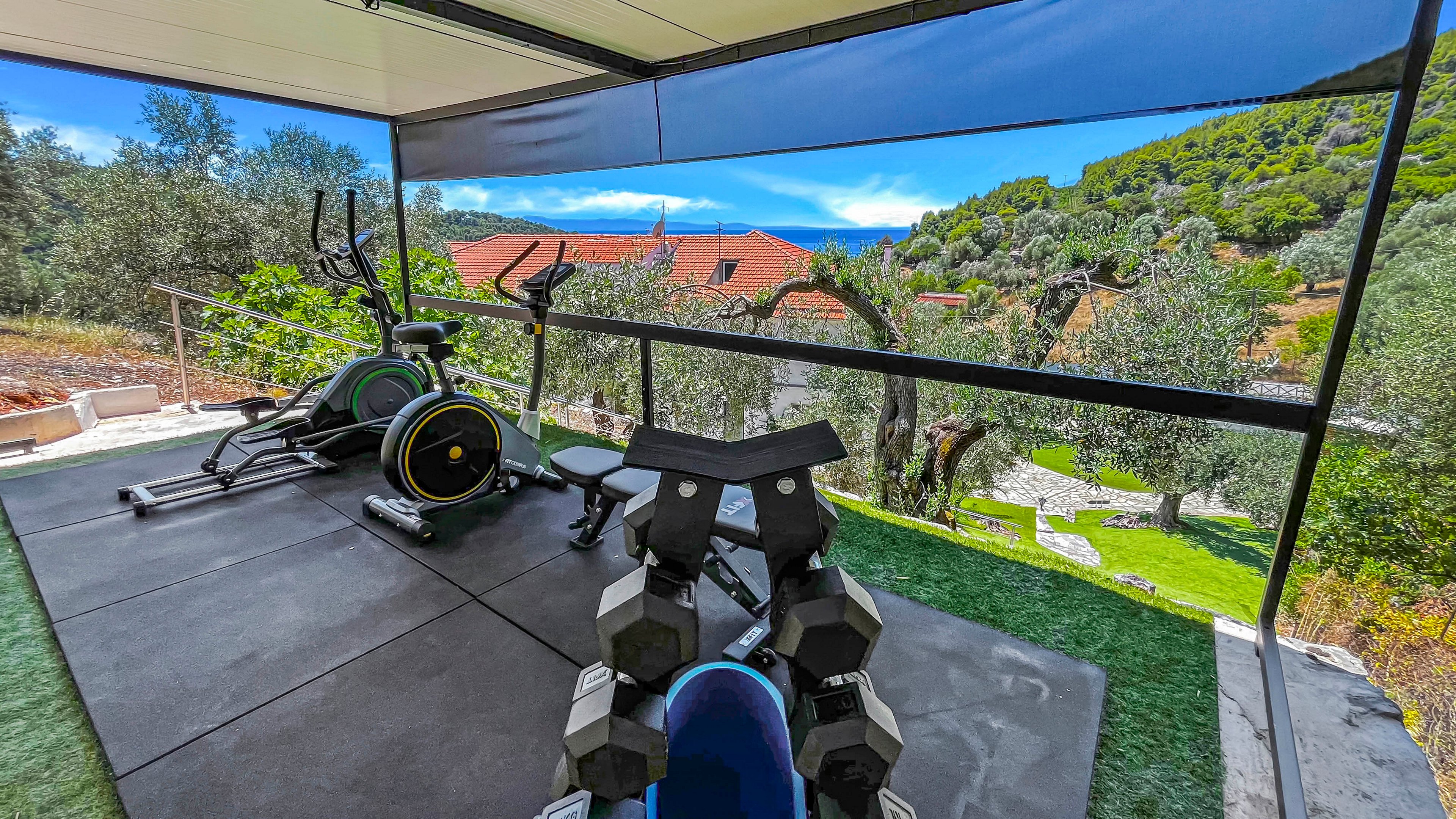 An open-air home gym is set up on a covered terrace overlooking a lush hillside landscape. The space features an elliptical trainer, a weight bench, and a rack of dumbbells arranged on rubber flooring. Glass or clear railings frame the view of red-roofed houses, trees, and distant hills under a bright blue sky. The scene combines fitness equipment with a tranquil, natural environment.