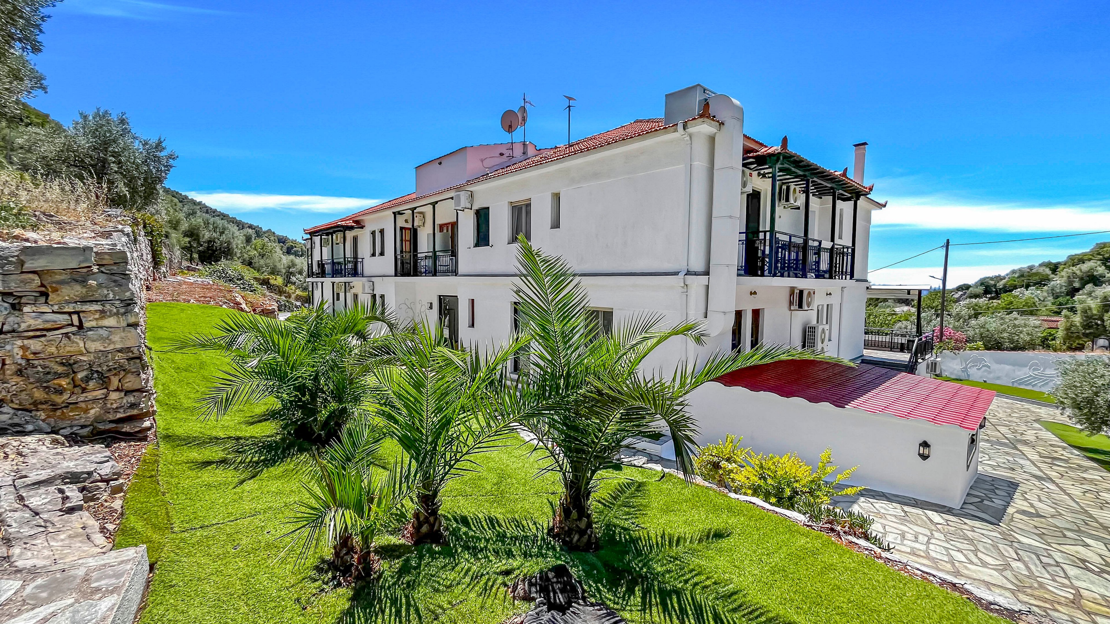 A white multi-story villa stands on a green hillside under a bright blue sky. The building features balconies with glass or dark railings and a red-roofed carport or covered area on the side. A neatly maintained lawn with several small palm trees and stone pathways surrounds the house. Rolling hills and scattered trees frame the property, suggesting a peaceful rural or coastal setting.