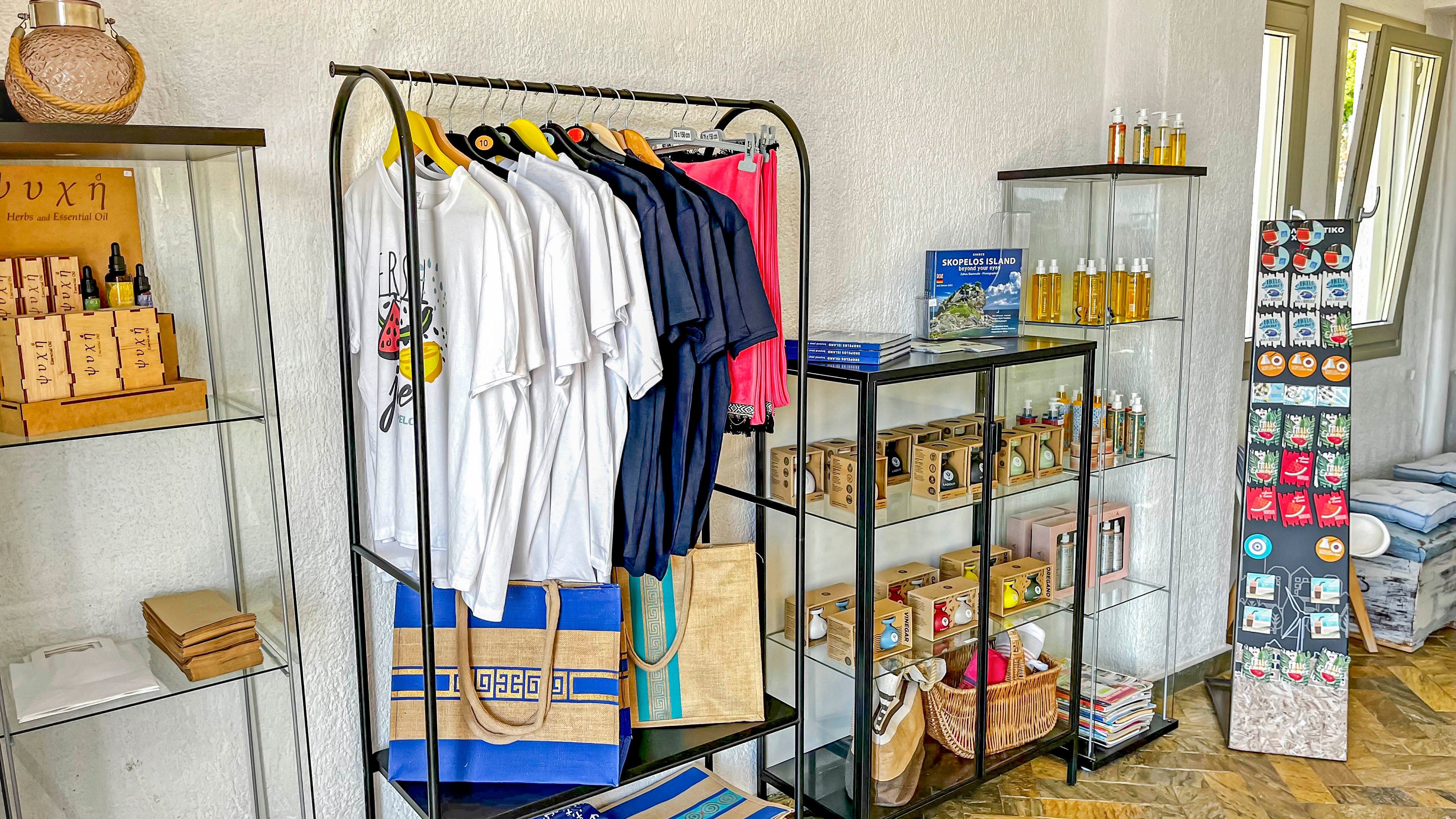 A small boutique retail space displays neatly arranged apparel and gift items. T-shirts in white, navy, and pink hang on a central rack above striped tote bags. Glass shelves hold boxed products, bottles, and woven baskets, while a tall stand near the window showcases magnets or small souvenirs. Natural light from the windows brightens the white walls and wooden floor, creating a clean and inviting atmosphere.