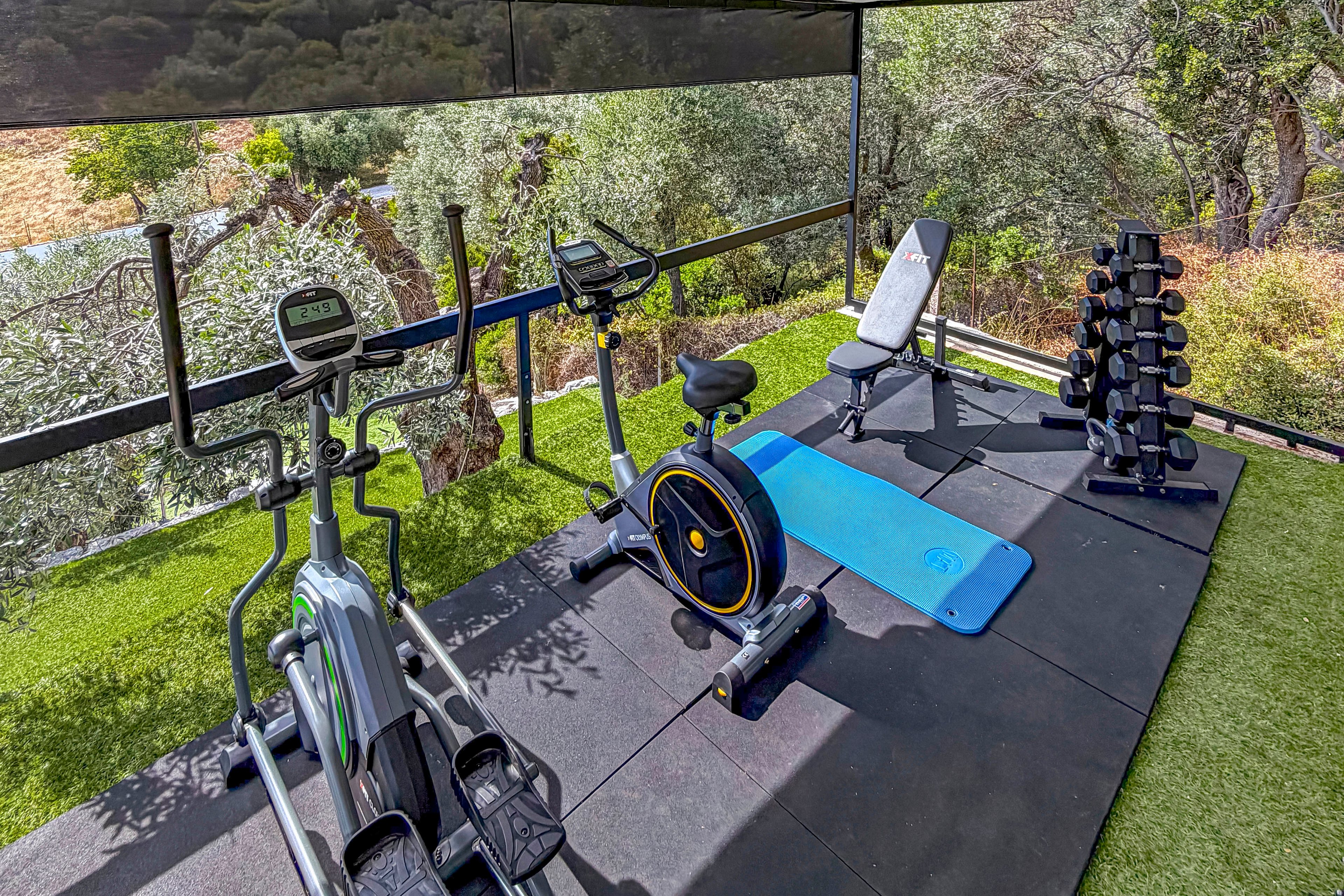 An outdoor home gym setup features exercise bikes, a weight bench, and a rack of dumbbells on rubber flooring. A blue yoga mat is laid out beside the bench, ready for stretching or floor exercises. The gym area is surrounded by green artificial turf and overlooks trees and natural scenery, creating a refreshing workout environment. The equipment is neatly arranged, suggesting a dedicated fitness space for strength and cardio training.