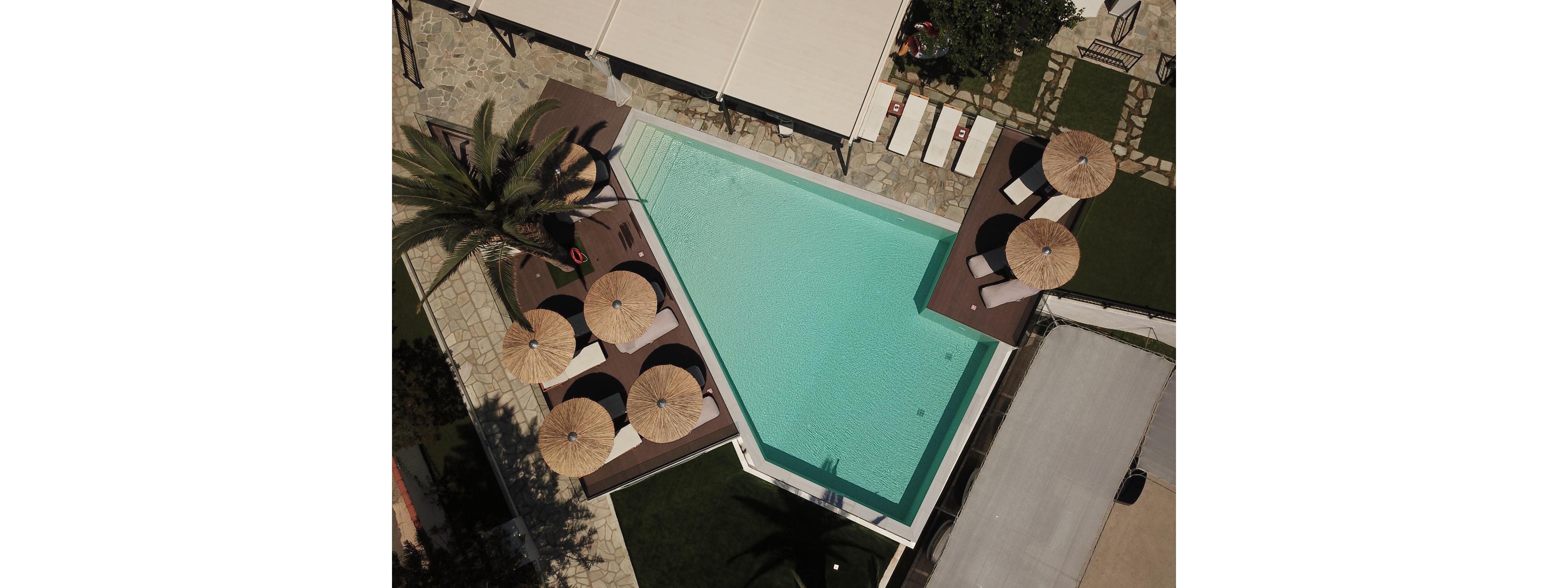 An aerial view shows a modern triangular swimming pool with clear turquoise water. Several round straw parasols and sun loungers are arranged neatly around the pool deck. Stone pathways, green lawn areas, and a few trees frame the poolside setting, creating a resort-like atmosphere. The scene appears calm and inviting, suggesting a relaxing vacation or hotel environment.