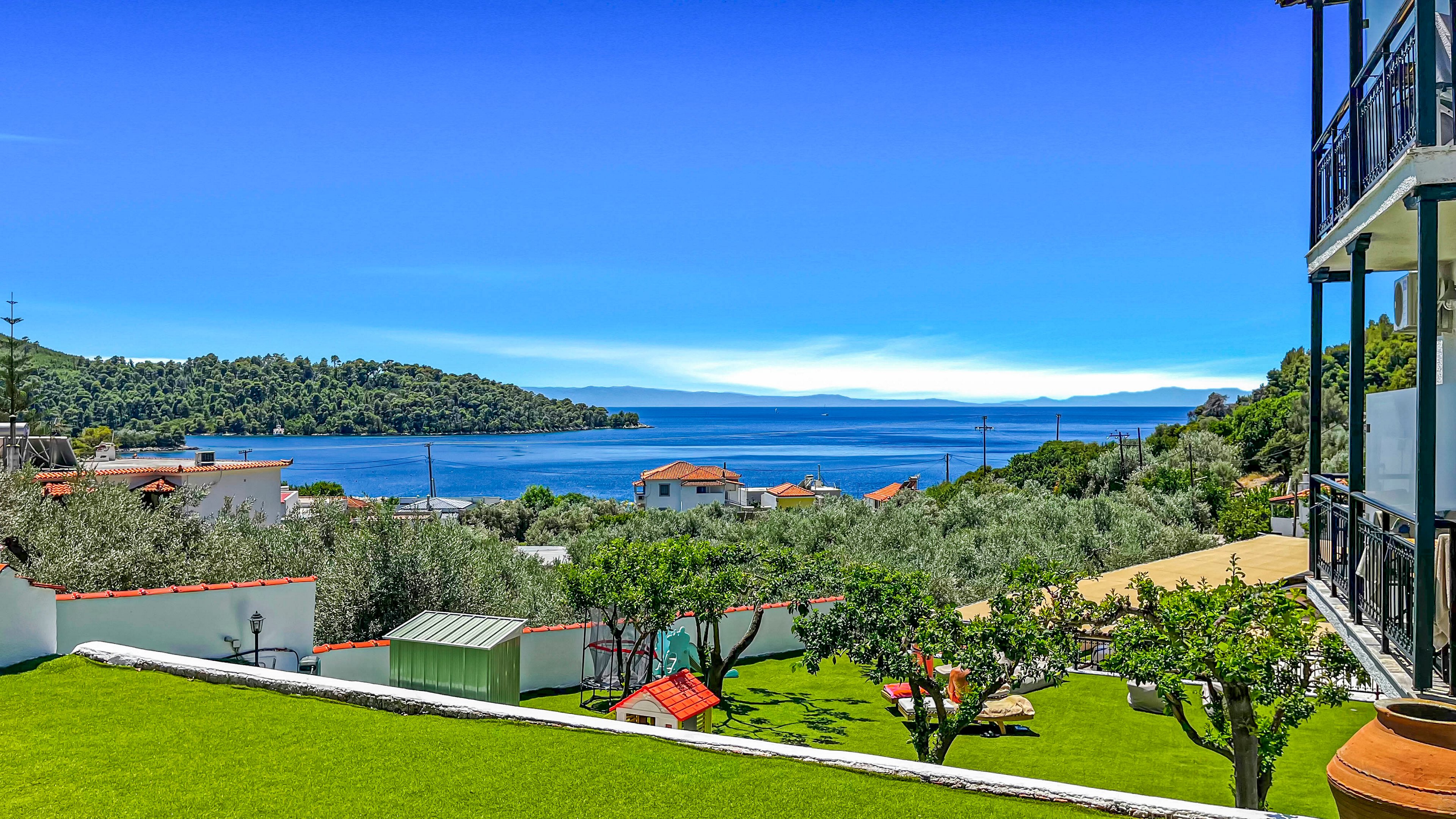 A vibrant coastal landscape features a neatly maintained green lawn and small garden structures on a hillside. In the middle distance, white houses with red-tiled roofs sit among trees and shrubs. Beyond the village, calm blue water stretches toward a forested peninsula and distant mountains under a clear sky. The scene is viewed from a balcony or terrace, suggesting a peaceful seaside retreat.