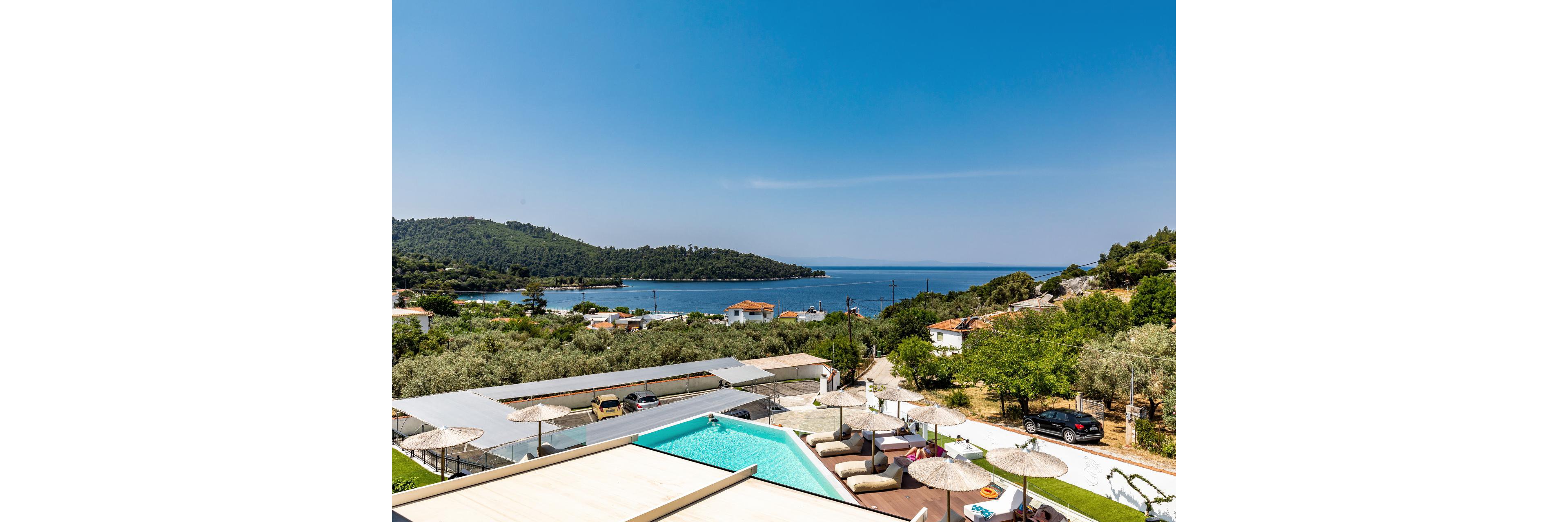 A modern hillside resort features a turquoise swimming pool with sun loungers and parasols in the foreground. Below the pool area, a small parking lot and buildings sit among lush green trees and olive groves. In the distance, a calm blue bay stretches out toward forested hills under a clear cloudless sky. The scene suggests a tranquil vacation destination with scenic sea and mountain views.