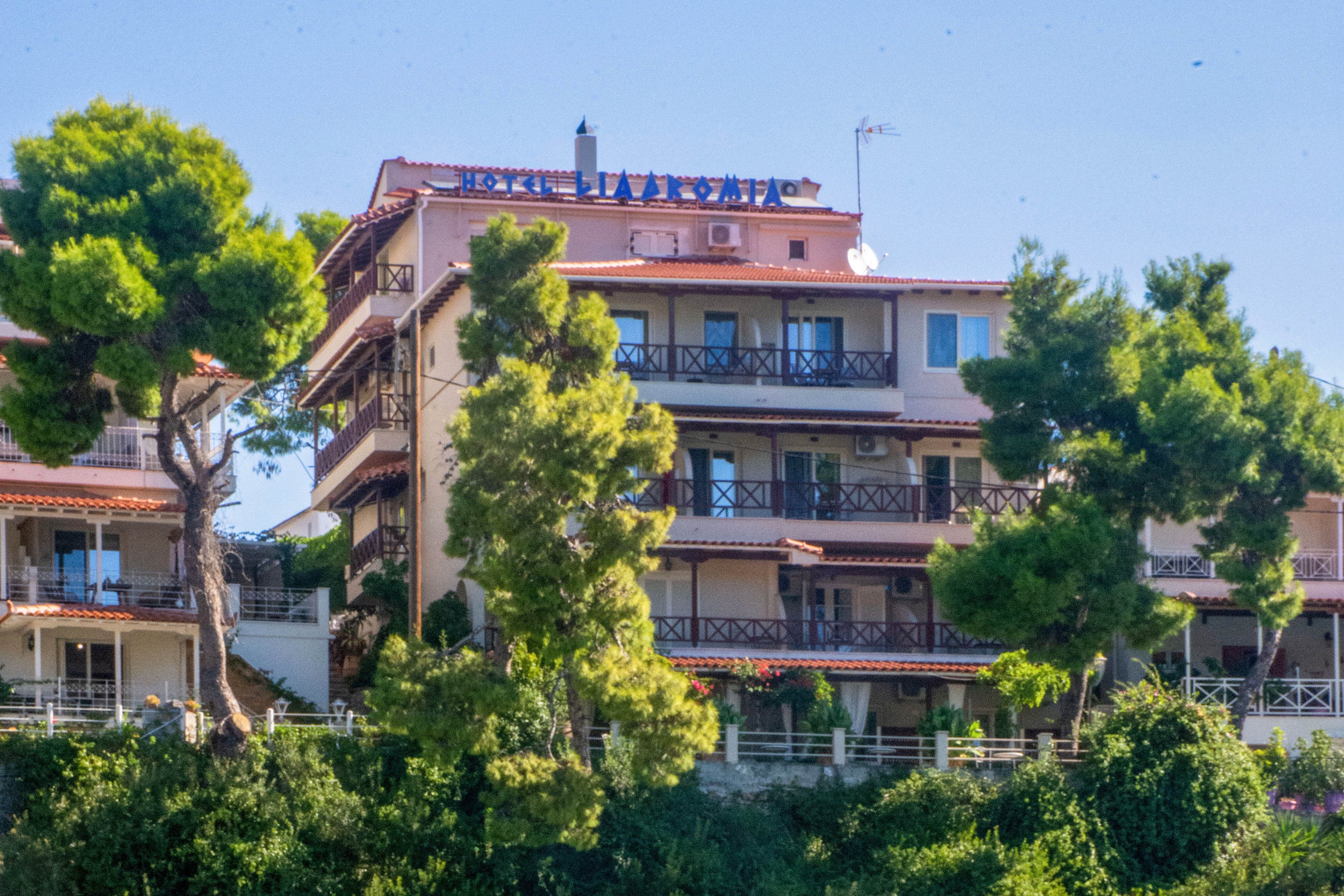 A multi-story seaside hotel stands on a lush, green hillside overlooking a small harbor. Several cars are parked along the waterfront road at the base of the cliff. Small boats float on the calm turquoise water in the foreground, tied to the quay. Tall trees and dense vegetation surround the buildings, creating a Mediterranean coastal atmosphere. The scene is bright and sunny with clear blue skies above.