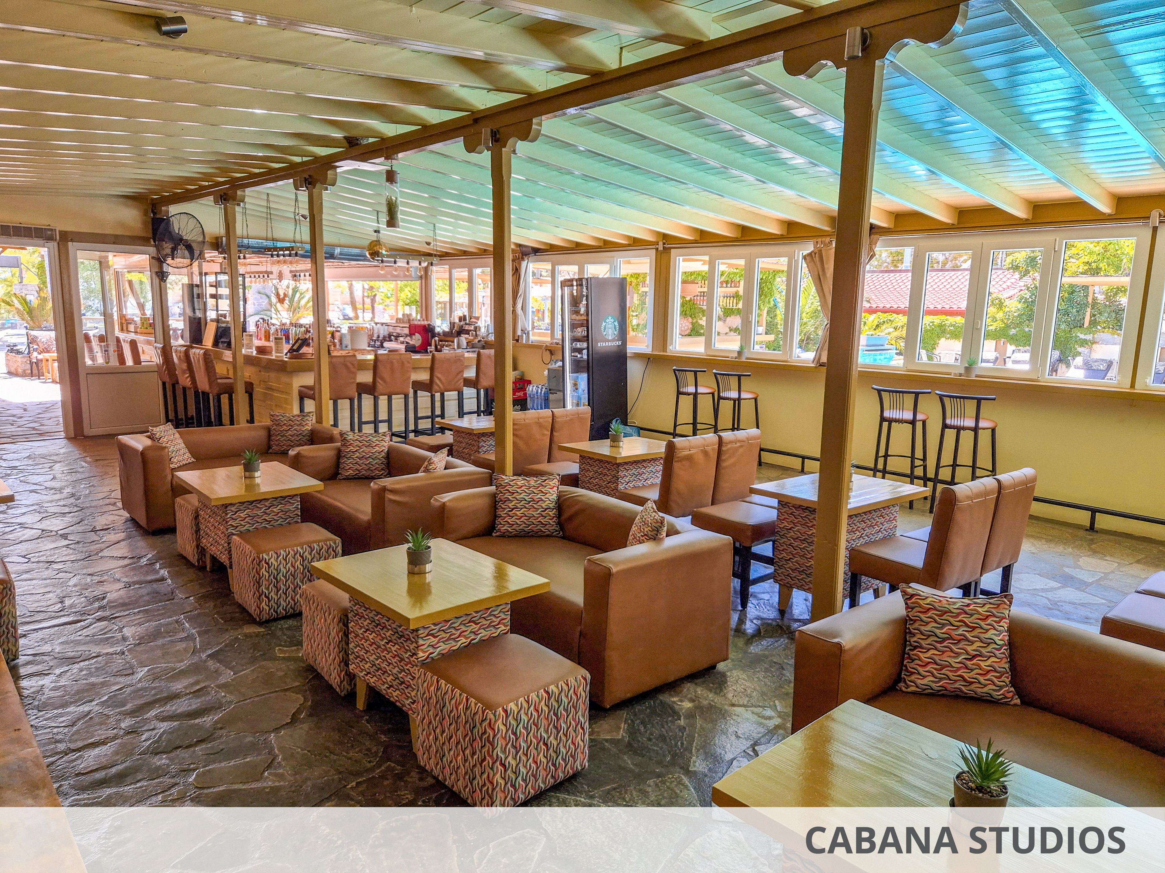 Spacious indoor lounge area featuring multiple low wooden tables and cushioned armchairs arranged in rows. The setting appears to be a café or bar with a long counter and high stools visible in the background. Large windows line the wall, letting in natural light and offering views of greenery outside. Colorful patterned cushions and small potted plants on the tables add a relaxed, inviting atmosphere.