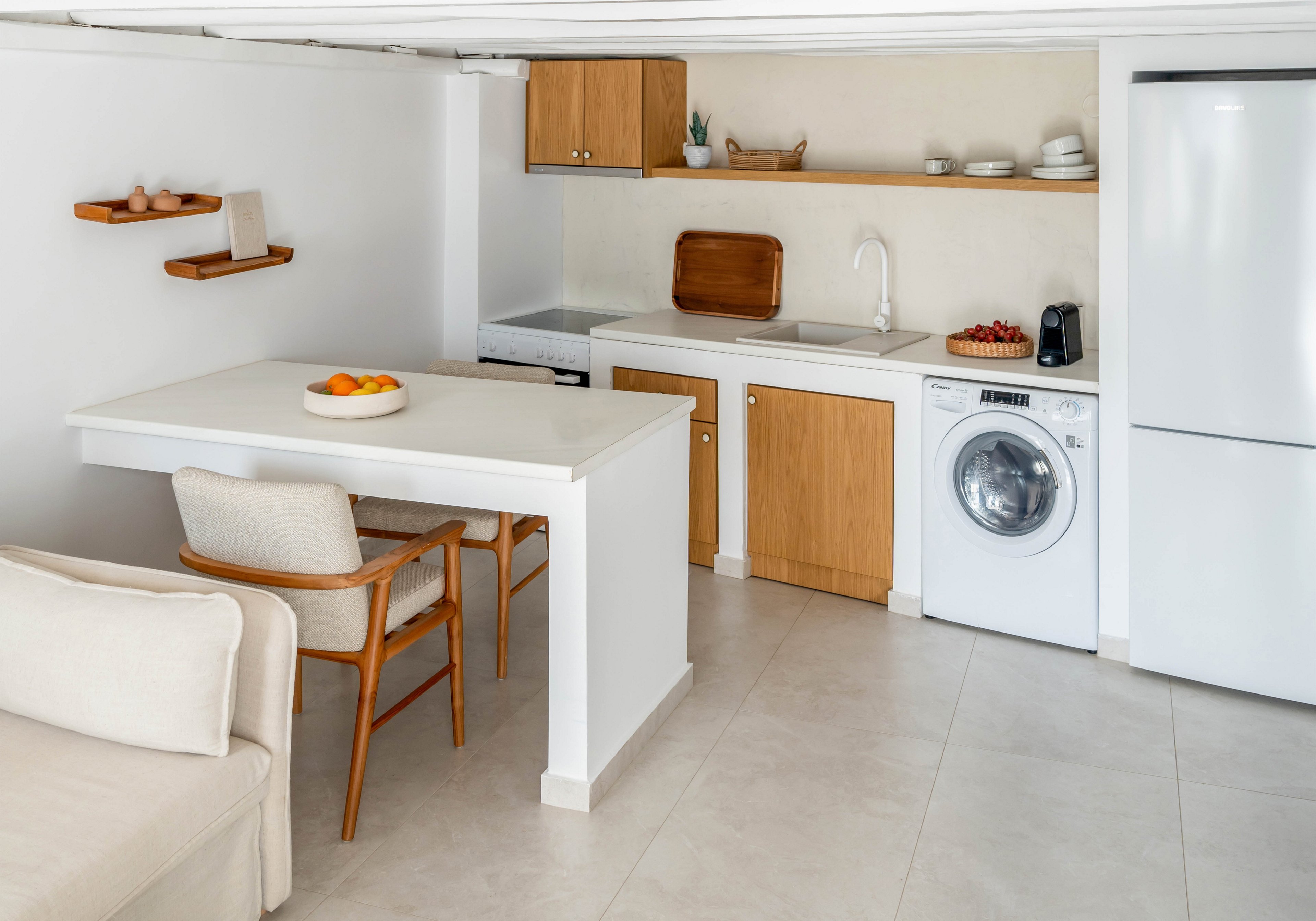 A small modern kitchen features white cabinetry with light wood accents and integrated appliances. A compact dining table with two wooden chairs extends from the counter near a beige sofa. Open shelves display dishes and decor, while a washing machine and tall refrigerator line one wall. The space is bright, tidy, and designed in a minimalist style with neutral tones.