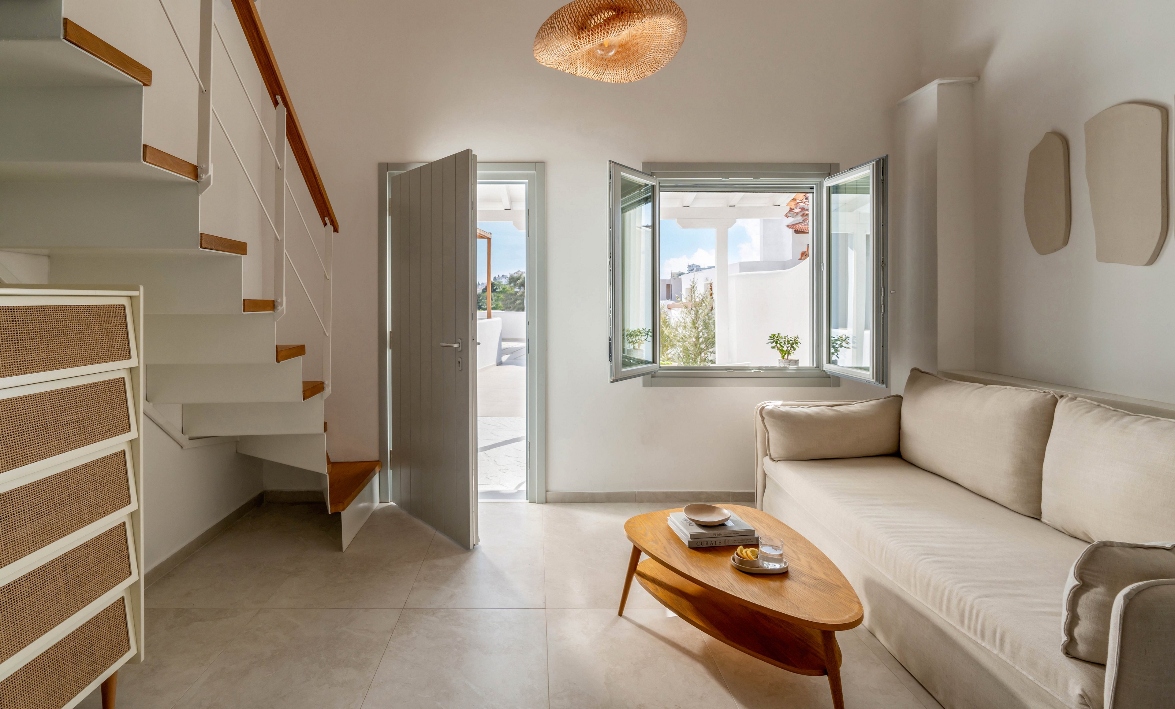 A modern living room features a light-colored sofa and a sleek wooden coffee table. An open staircase with white steps and wooden treads rises along the left wall. Sunlight streams through open windows and a glass door, revealing a bright outdoor terrace. Neutral tones, clean lines, and minimal decor create an airy, contemporary atmosphere.