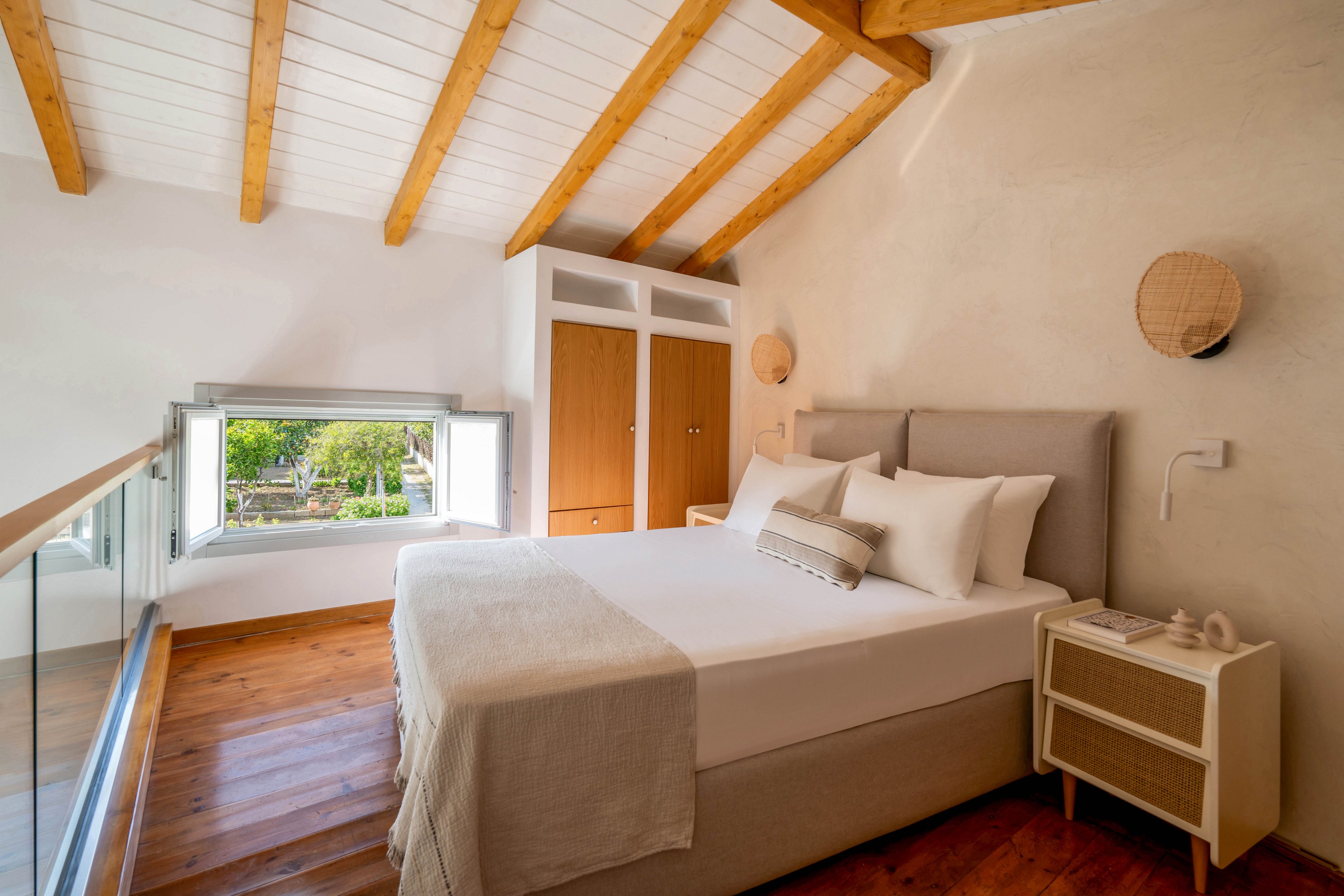 A cozy attic bedroom features a neatly made double bed with neutral linens and multiple pillows. The space has sloped white ceilings with exposed wooden beams and warm wooden flooring. A built-in wardrobe with white framing and wooden doors stands at the far end of the room. Natural light floods in through a low, open window overlooking greenery, enhancing the airy and minimalist atmosphere.