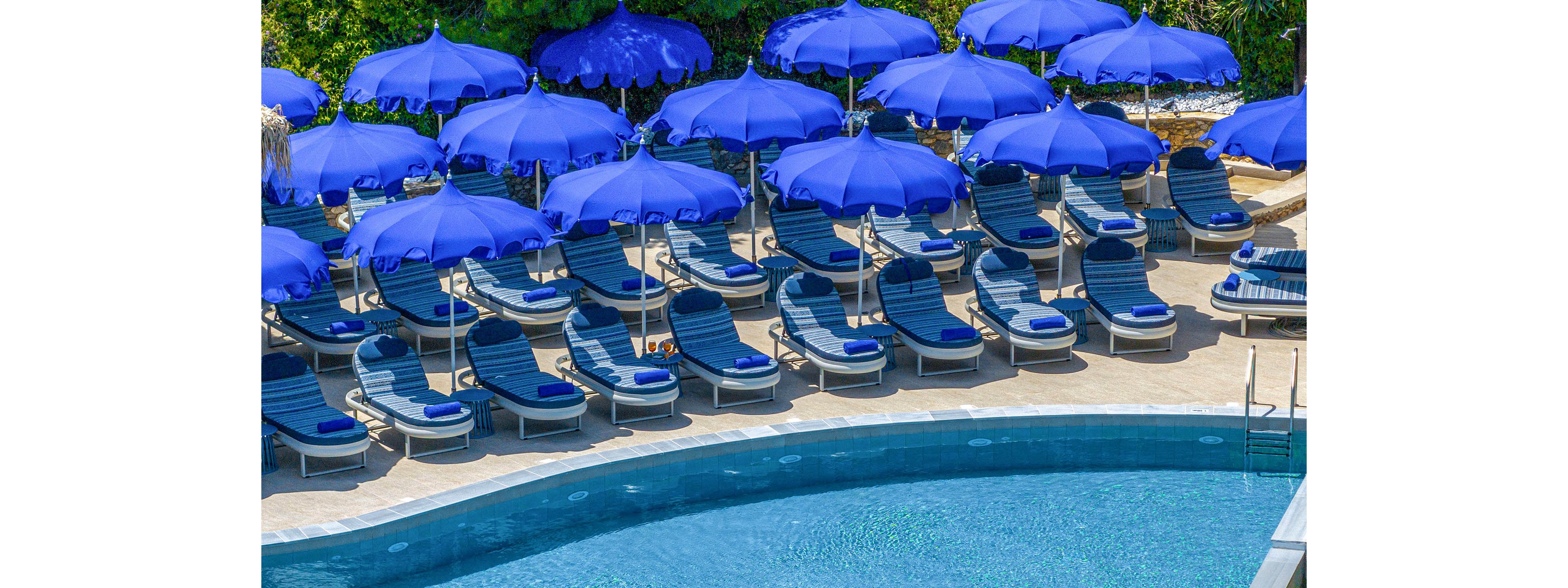 A curved swimming pool sits in the foreground of a sunny resort scene. Behind it, multiple rows of empty lounge chairs are neatly arranged under bright blue umbrellas. The pool water appears clear and inviting, reflecting the vivid colors around it. Lush greenery in the background adds a tropical feel to the tranquil, vacation setting.