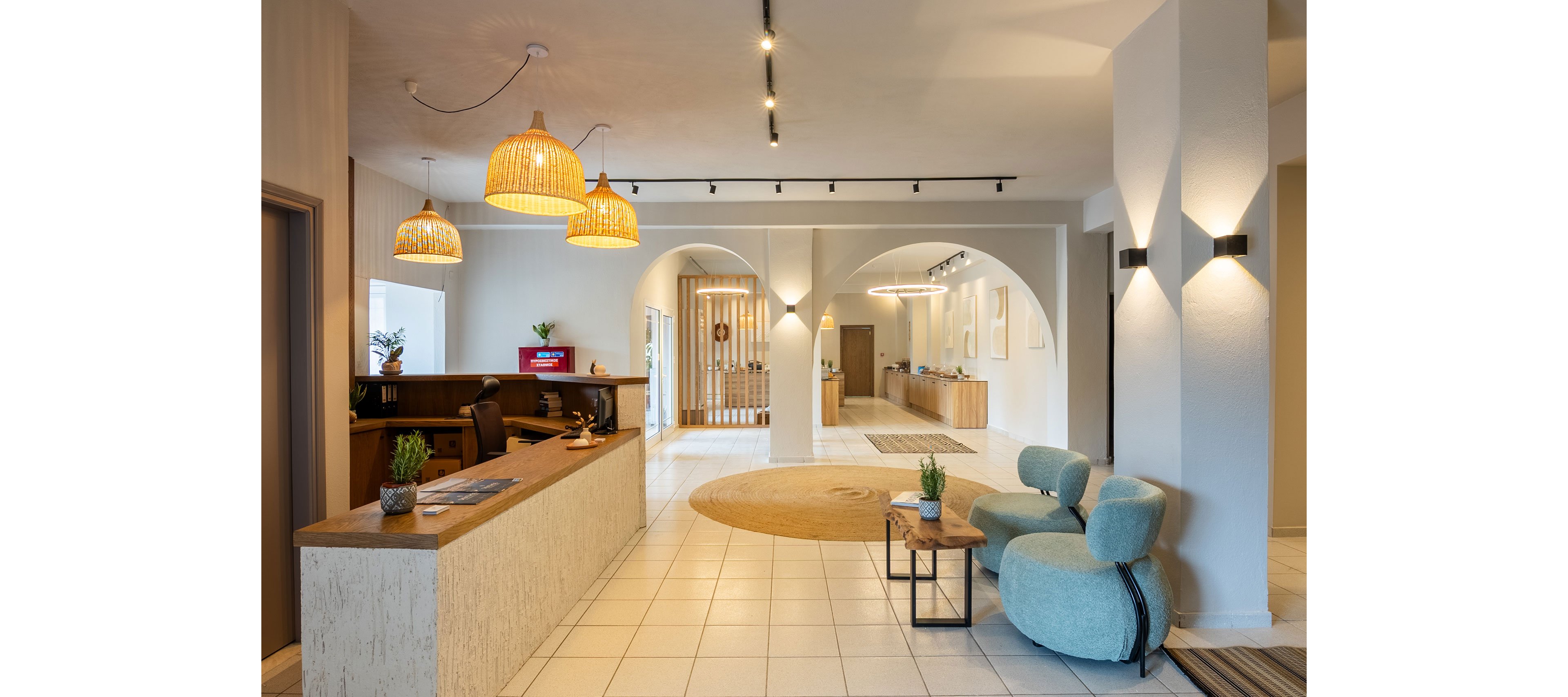 A contemporary hotel lobby features a wooden reception desk on the left and a cozy seating area with rounded blue chairs on the right. Warm pendant lights hang from the ceiling, illuminating the tiled floor and neutral-colored walls. Arched openings lead to a hallway with additional wooden furniture and framed artwork. Small potted plants and decorative objects add a welcoming, minimalist touch to the interior.