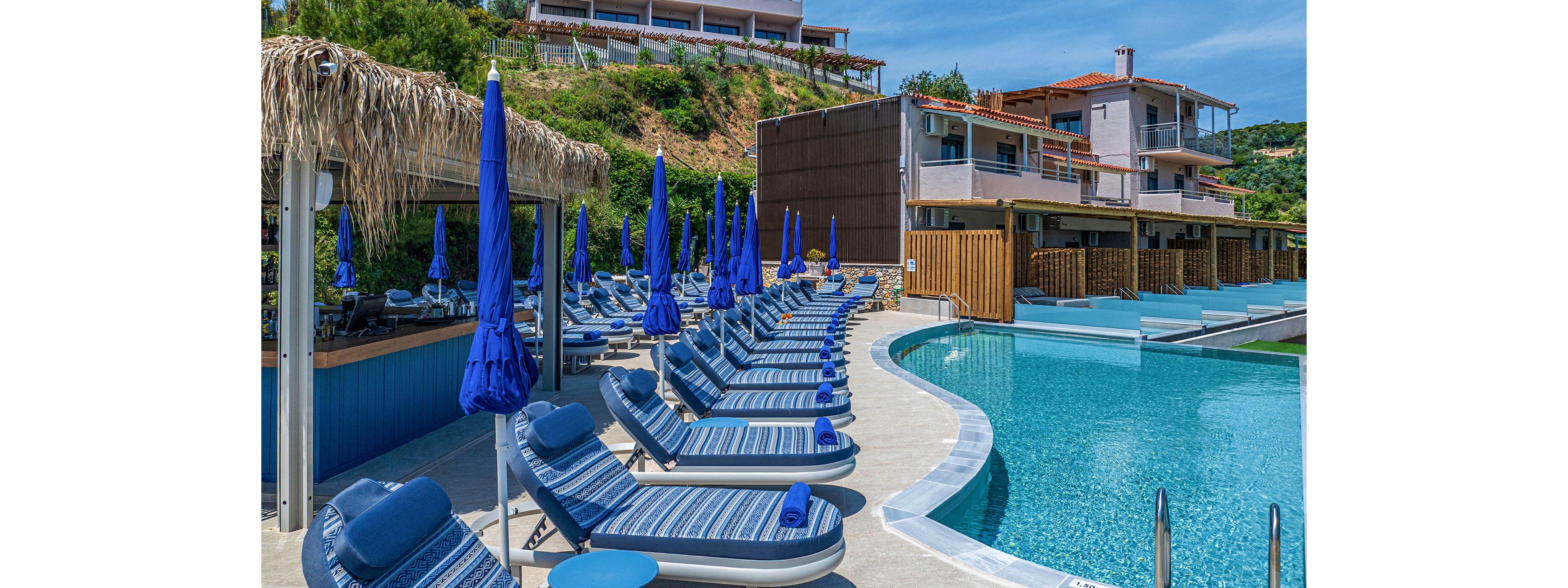 A modern resort infinity pool is lined with rows of blue cushioned sun loungers and matching umbrellas. The pool curves alongside a contemporary hotel building with balconies and wooden accents. Lush green hills and additional terraced structures rise in the background under a clear blue sky. The scene suggests a sunny, relaxing vacation atmosphere at a coastal or hillside retreat.