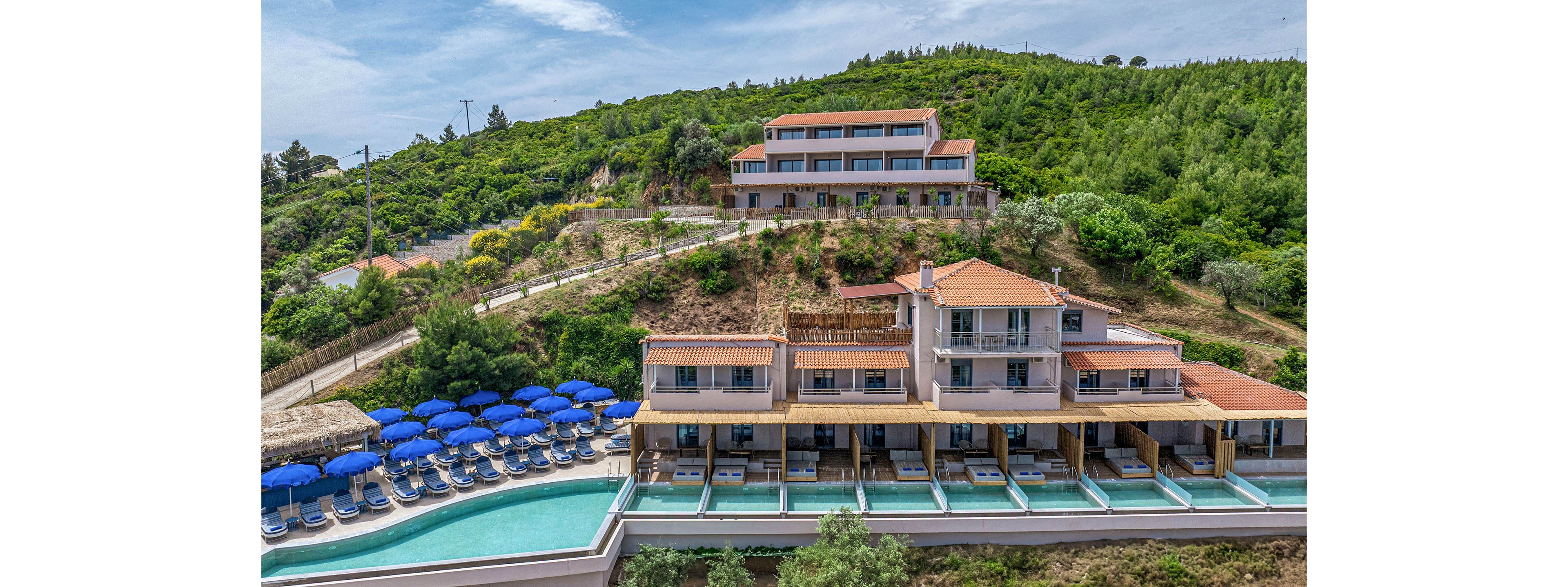 A multi-level resort complex is built on a lush green hillside overlooking a long swimming pool. Rows of blue sun umbrellas and loungers line the poolside terrace, suggesting a relaxing vacation setting. The buildings feature terracotta roofs and balconies facing the water, emphasizing Mediterranean-style architecture. Dense trees and vegetation surround the property, creating a secluded and tranquil atmosphere.