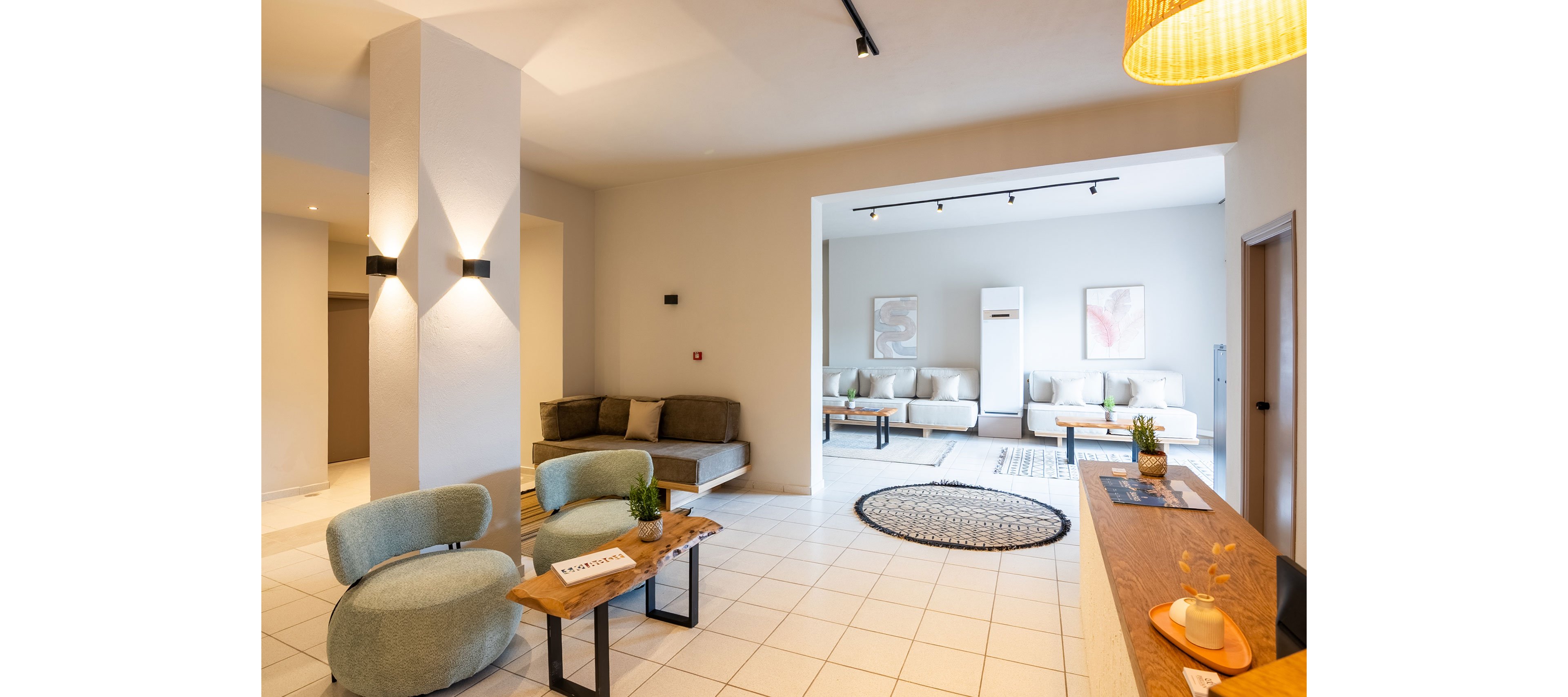 Bright contemporary lounge area featuring modern sofas and armchairs in neutral tones. The space is divided into two seating zones with tiled flooring and soft, indirect lighting. A wooden console and coffee table display small plants and decorative objects, adding warmth to the minimalist design. Abstract wall art and a round woven rug complete the cozy, welcoming atmosphere.