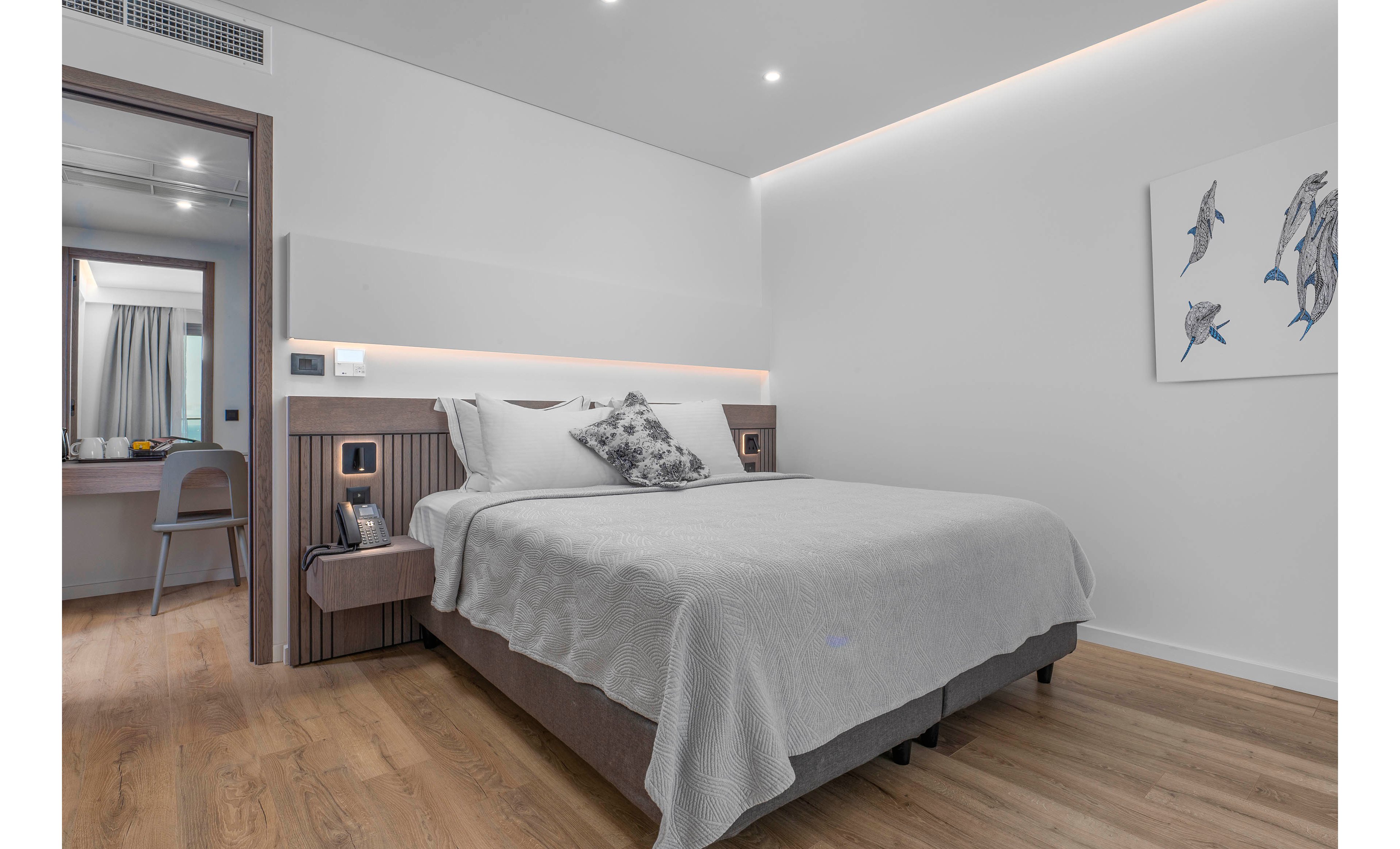 A modern hotel bedroom features a neatly made double bed with light gray bedding and decorative pillows. The room has a minimalist design with clean white walls, wooden flooring, and built-in bedside lighting. A small desk and chair are visible in the adjoining vanity area with a mirror and curtain. A framed artwork of fish adds a subtle decorative touch to the otherwise neutral space.