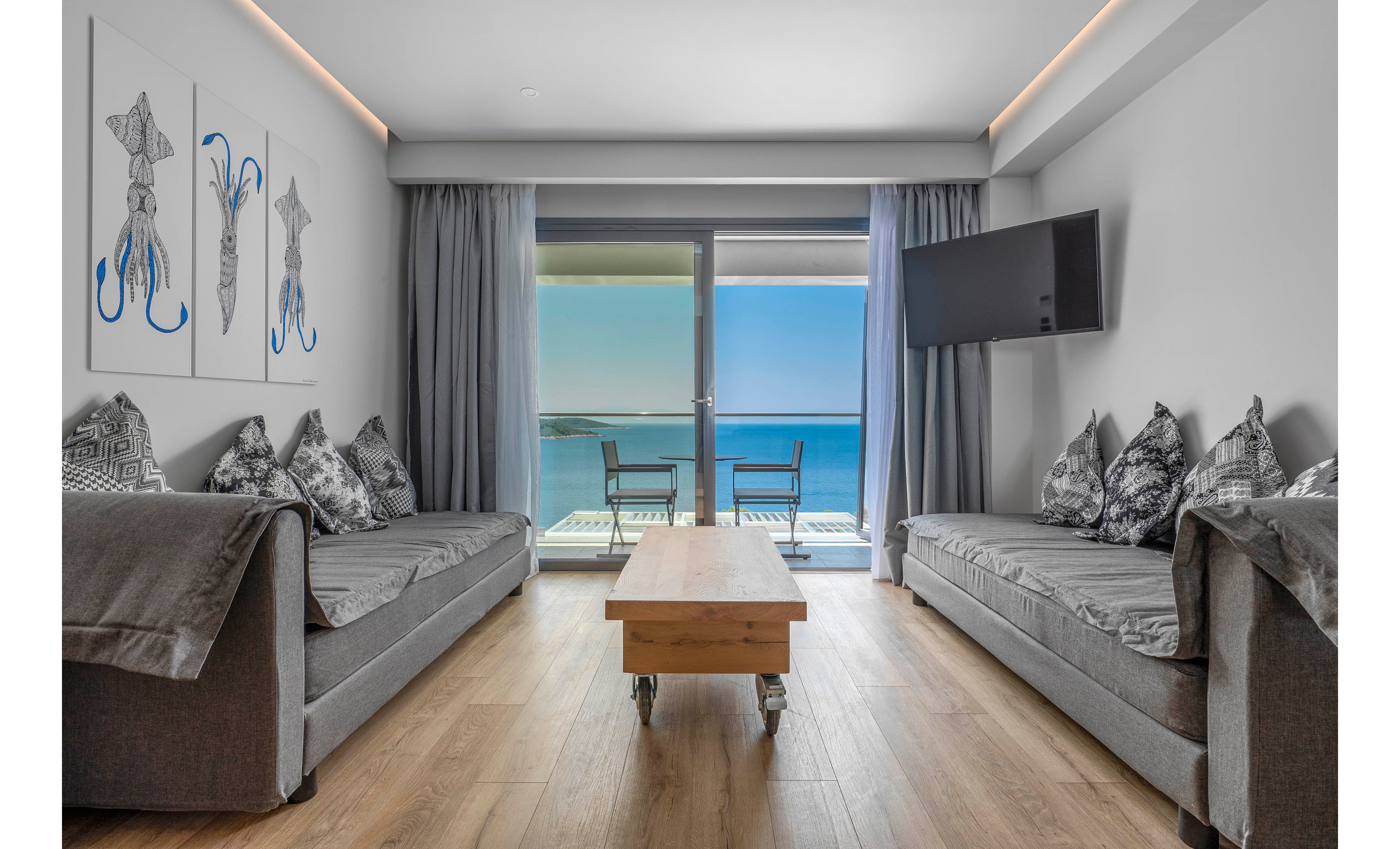 A contemporary living room features two grey sofas facing each other with a wooden coffee table on wheels between them. Floor-to-ceiling glass doors open onto a balcony with two chairs and a small table overlooking a bright blue ocean. Light grey curtains frame the view, and a wall-mounted flat-screen television is angled toward the seating area. Minimalist artwork of squid hangs on the wall, complementing the neutral tones and clean, modern design.