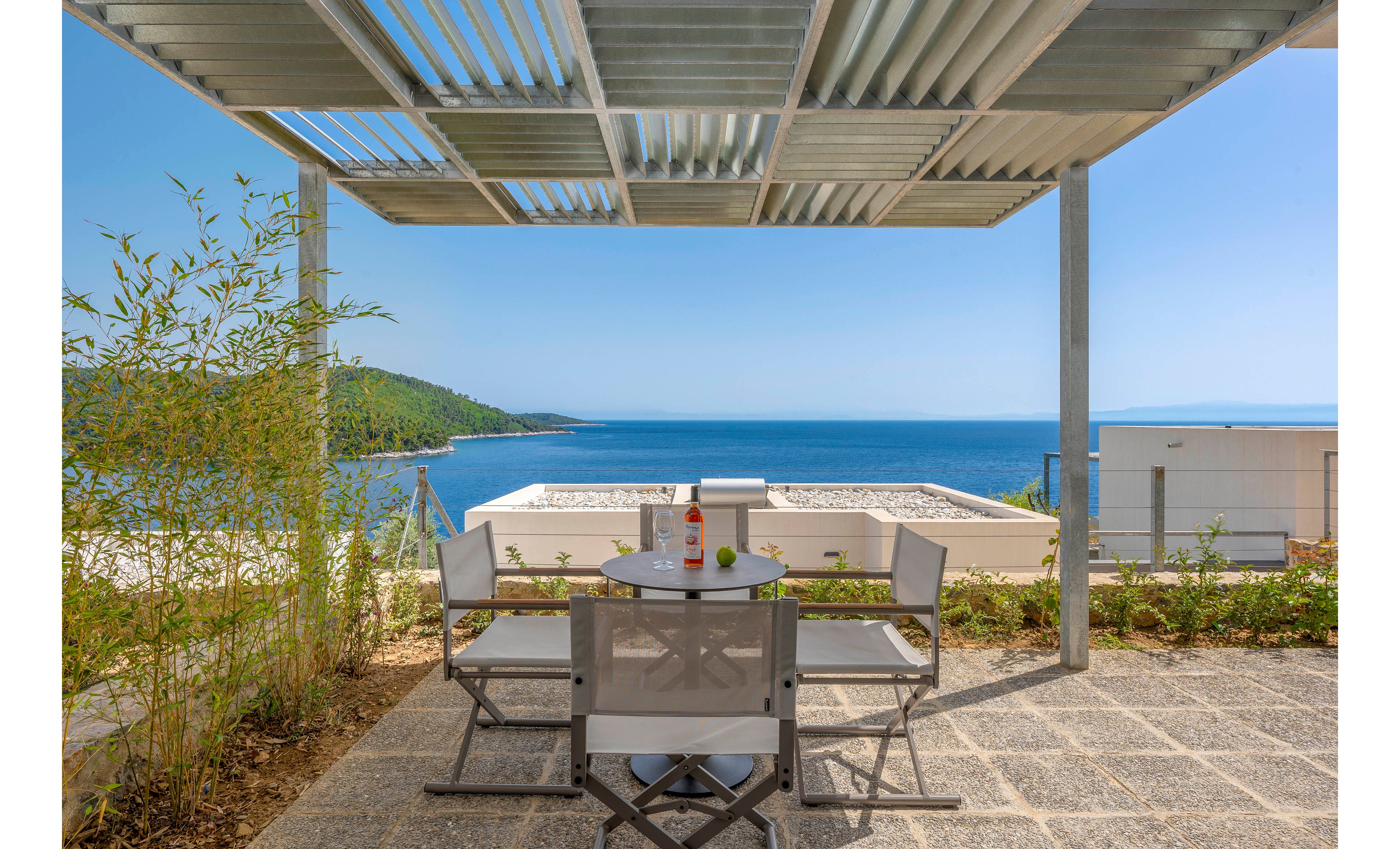A modern terrace dining area features a small round table with four chairs under a metal pergola. The terrace overlooks a bright blue sea and a distant green hillside, creating a serene coastal setting. Contemporary buildings and low walls frame the view while plants and bamboo add natural accents. Clear skies and calm water emphasize a relaxed, vacation-like atmosphere.