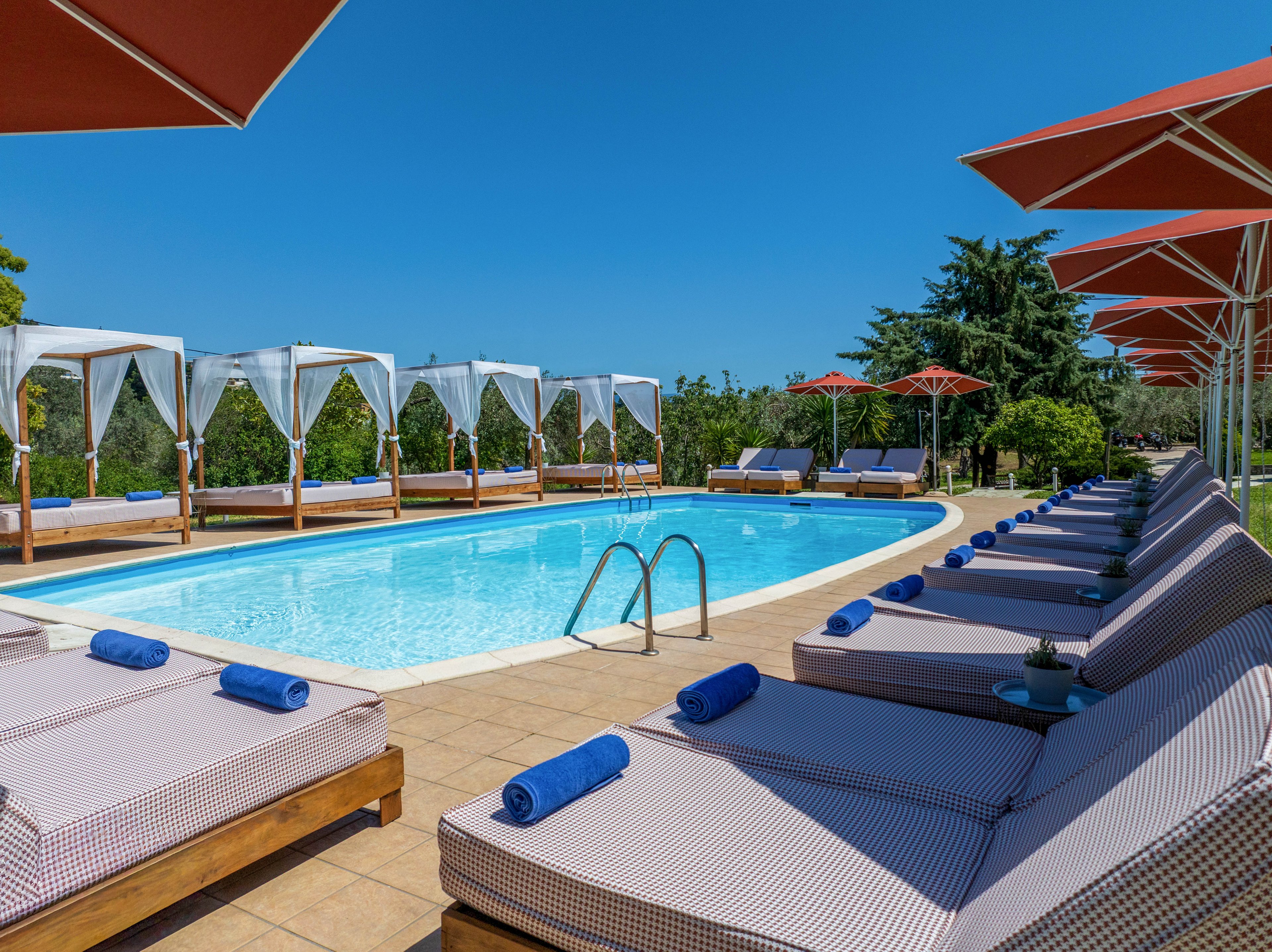 An outdoor swimming pool is surrounded by cushioned sun loungers with neatly rolled blue towels. White cabanas line one side of the pool, while large red umbrellas provide shade along the deck. The scene is set under a clear blue sky with lush green trees in the background. The overall atmosphere suggests a tranquil, upscale resort ideal for relaxation and sunbathing.