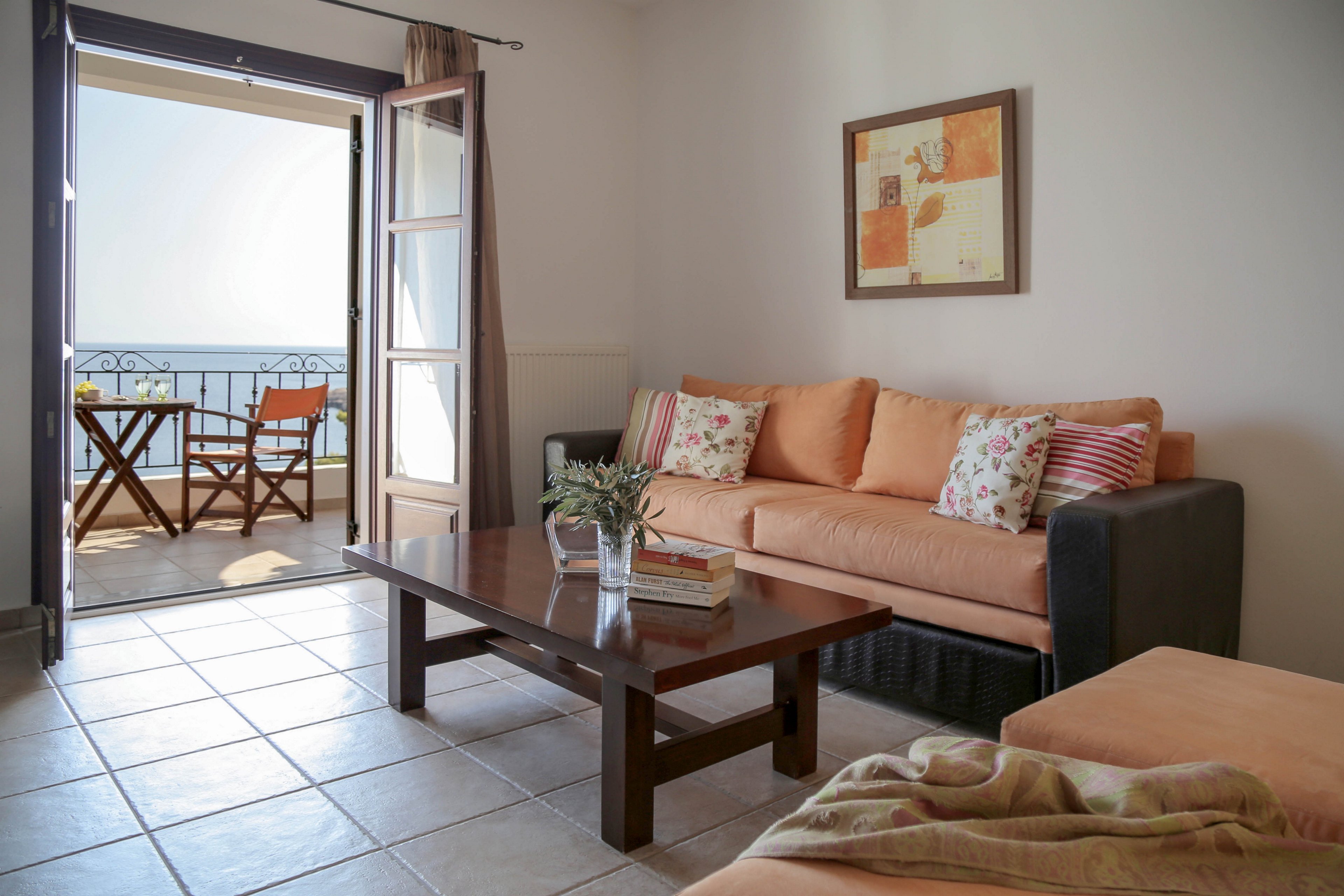 A cozy living room features a peach-colored sofa with patterned cushions and a dark wooden coffee table. Sunlight streams in through open French doors leading to a small balcony with a table and two chairs. The tiled floor and neutral walls create a clean, airy feel, complemented by a framed artwork above the sofa. Beyond the balcony, a calm sea and clear sky suggest a relaxing coastal setting.