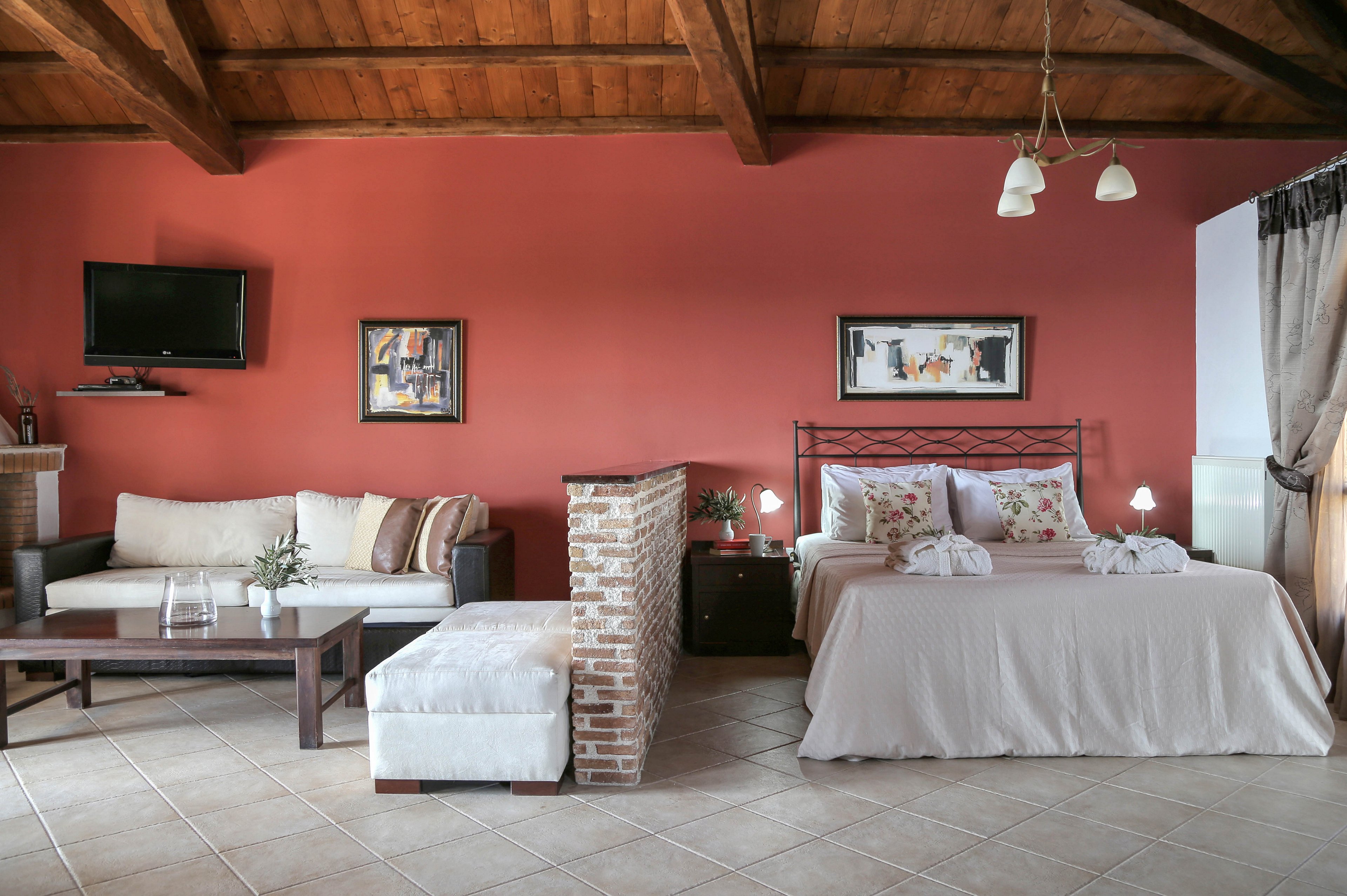 A cozy studio-style bedroom features a large bed with neatly folded towels shaped decoratively on top. The room combines a sleeping area and a sitting area with a sofa, coffee table, and upholstered bench separated by a low brick partition. A red accent wall, wooden ceiling, and tiled floor create a warm, rustic atmosphere. Wall art, bedside lamps, and a mounted television add decorative and functional touches to the space.