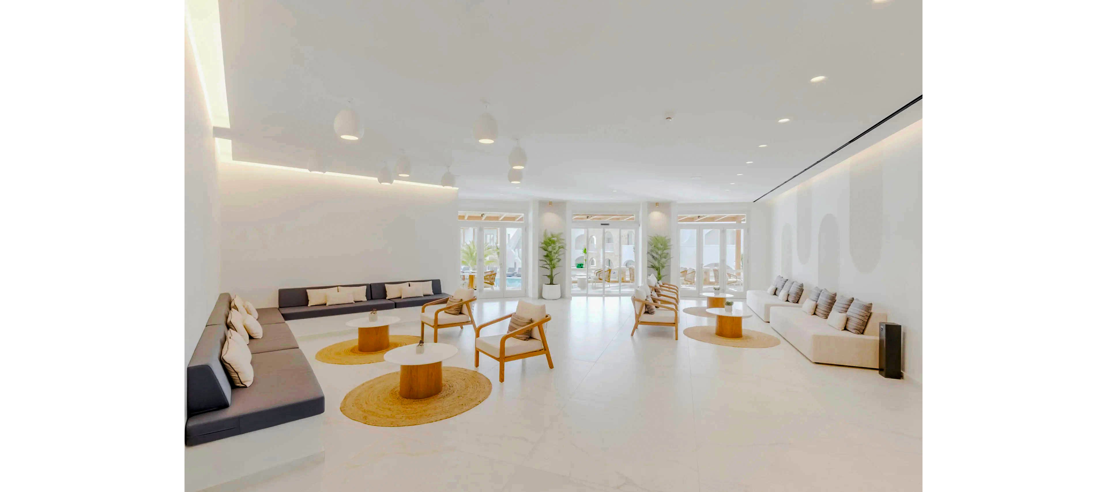 Spacious contemporary lounge area featuring minimalist sofas and wooden chairs arranged around low circular tables. The room is predominantly white with light wood accents and neutral cushions. Large floor-to-ceiling windows at the far end let in abundant natural light and reveal an outdoor terrace. Recessed ceiling lights and pendant fixtures enhance the clean, airy atmosphere of the interior.