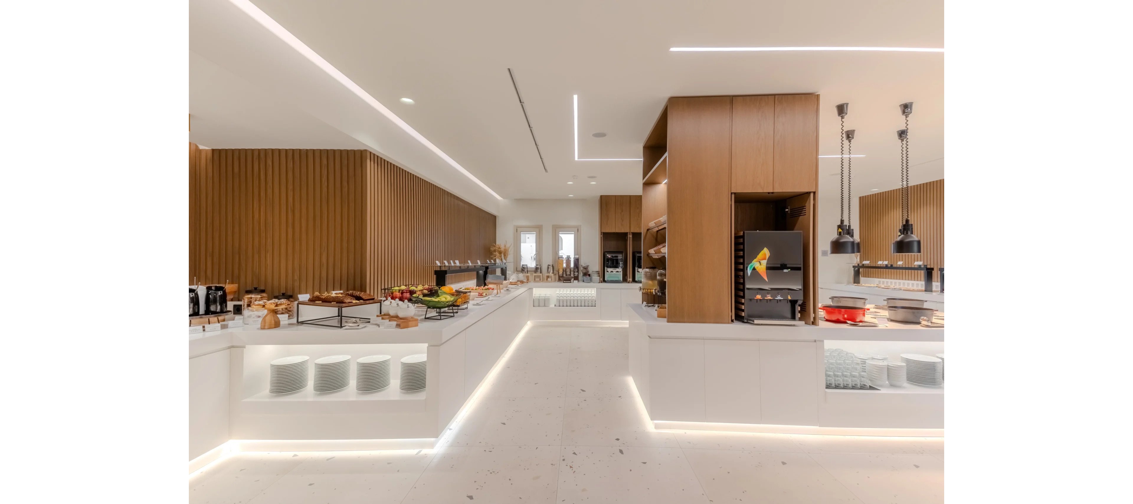 A spacious self-service buffet area features long white counters filled with breakfast items and beverages. Warm wooden wall panels and cabinetry contrast with the bright white floor and countertops. Recessed ceiling lights and under-counter LED strips create a clean, contemporary atmosphere. A drink dispenser and heated serving lamps add to the professional food service setup.