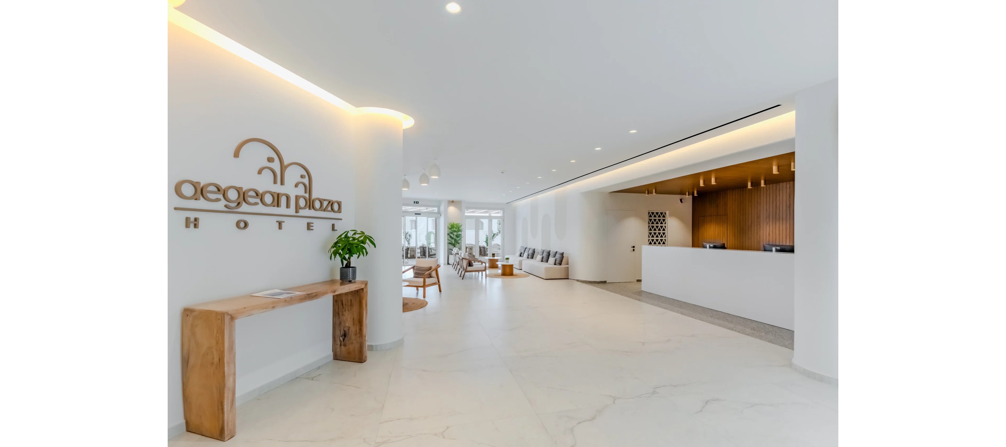 Spacious modern hotel lobby with a minimalist white interior and light wood accents. The Aegean Plaza Hotel name is displayed on the left wall above a simple wooden console table with a potted plant. A sleek white reception desk with warm wooden backdrop stands on the right, while lounge seating and large windows appear in the background. Soft recessed lighting and clean marble-style flooring create an airy, welcoming atmosphere.
