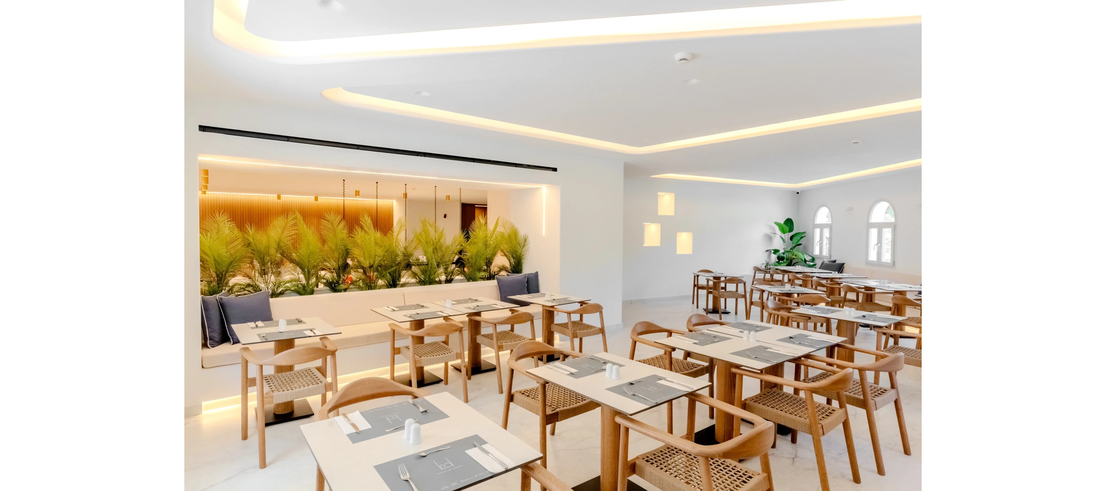 A contemporary restaurant dining area features light wooden tables and chairs neatly arranged in rows. The space is bright and minimalistic, with white walls, large windows, and recessed ceiling lighting. A built-in bench with cushions runs along one wall, backed by a row of green plants for a natural accent. Table settings are simple and clean, creating an inviting atmosphere for guests.