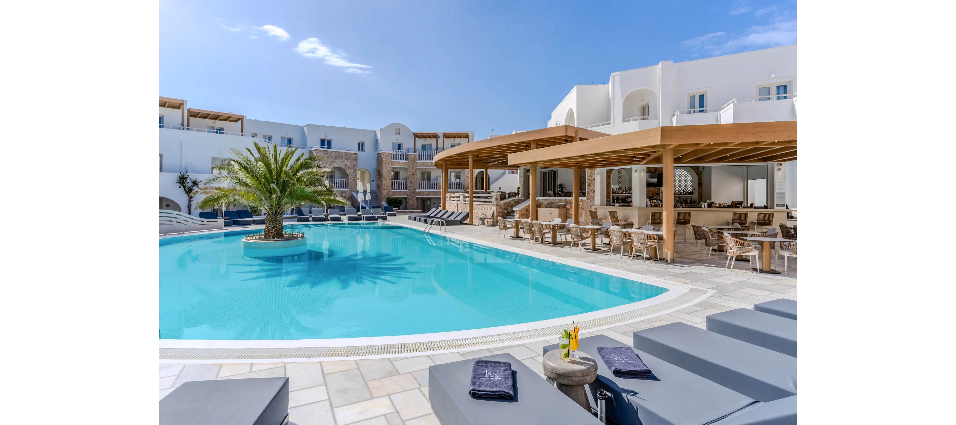 A luxurious resort pool area features a large turquoise swimming pool surrounded by modern white buildings. A single palm tree stands on an island in the middle of the water, adding a tropical touch. Comfortable gray sun loungers with towels and drinks are arranged along the poolside, facing the water. A shaded wooden pergola shelters an open-air bar and dining area, creating a relaxed vacation atmosphere under a clear blue sky.