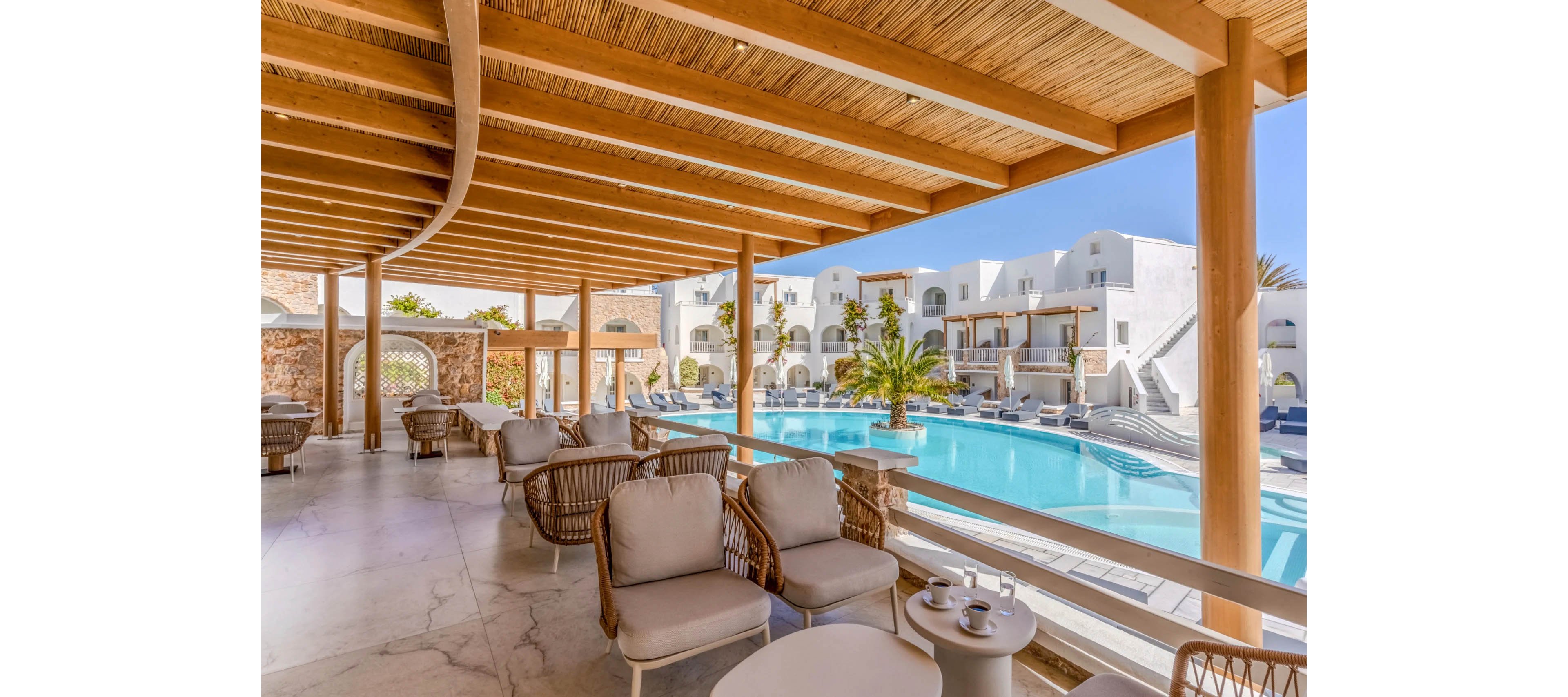 An elegant resort terrace with cushioned lounge chairs overlooks a large outdoor swimming pool. White, Mediterranean-style buildings with balconies surround the pool area under a clear sky. Wooden beams and a woven ceiling create a shaded seating space with tables set for coffee. Palm trees and sun loungers enhance the relaxed vacation atmosphere.