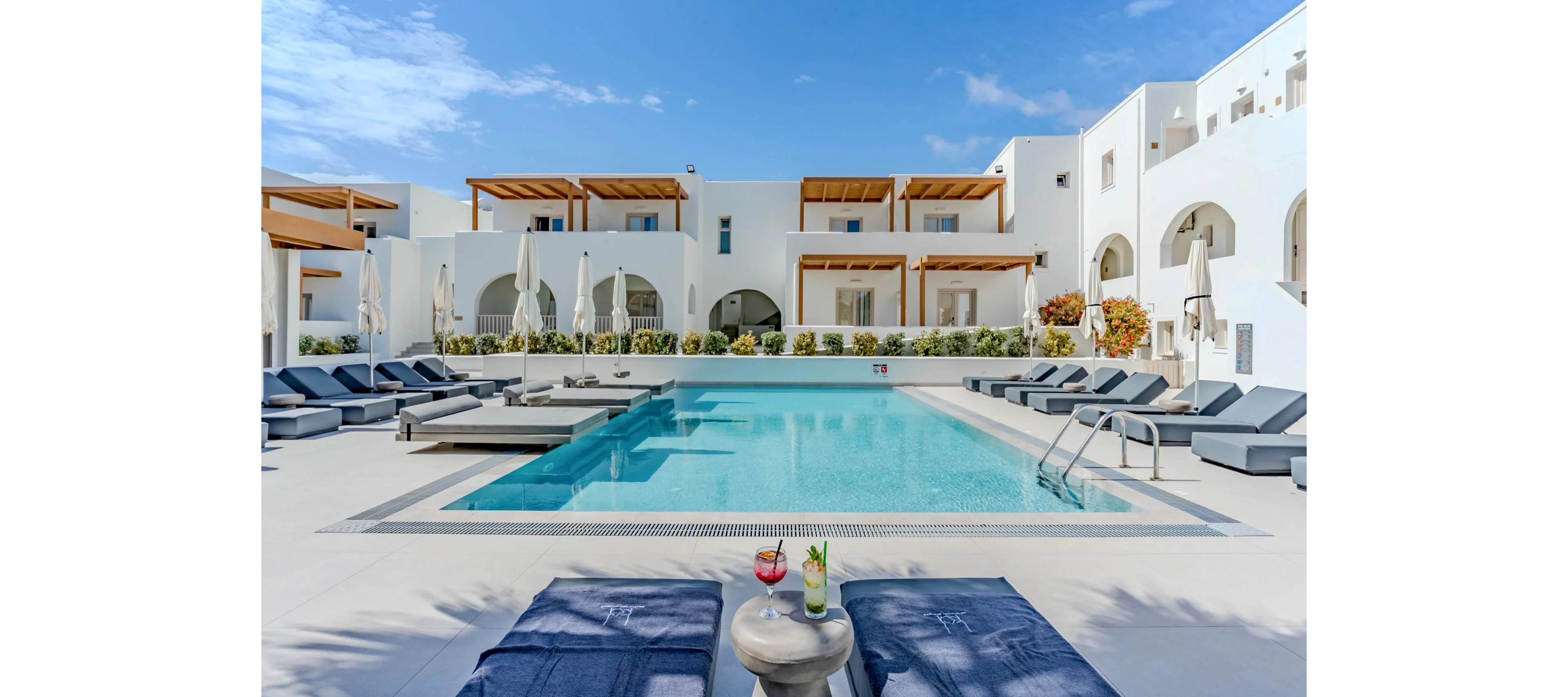 A serene outdoor swimming pool is centered in a modern white hotel courtyard. Sun loungers and closed umbrellas line both sides of the pool under wooden pergolas. In the foreground, two colorful cocktails rest on a small round table between towels on loungers. The bright blue sky and clear water create a relaxing vacation atmosphere. No people are visible, emphasizing calm and tranquility.
