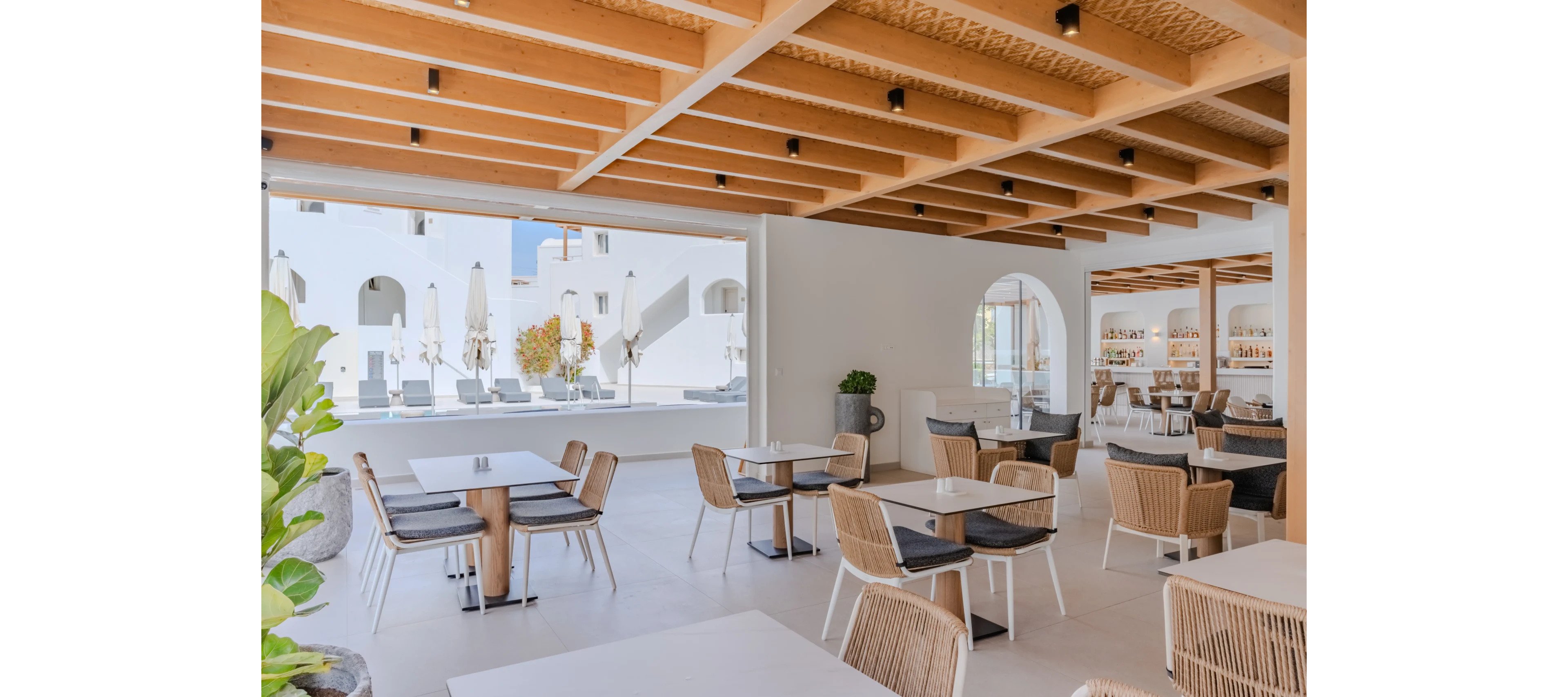 A contemporary open-plan restaurant features light wooden furniture and a geometric wooden ceiling. Neutral tones and minimalist decor create an airy, sophisticated dining space. Large openings frame views of a white resort courtyard with sun loungers and umbrellas. The setting appears calm and inviting, ideal for relaxed vacation dining.