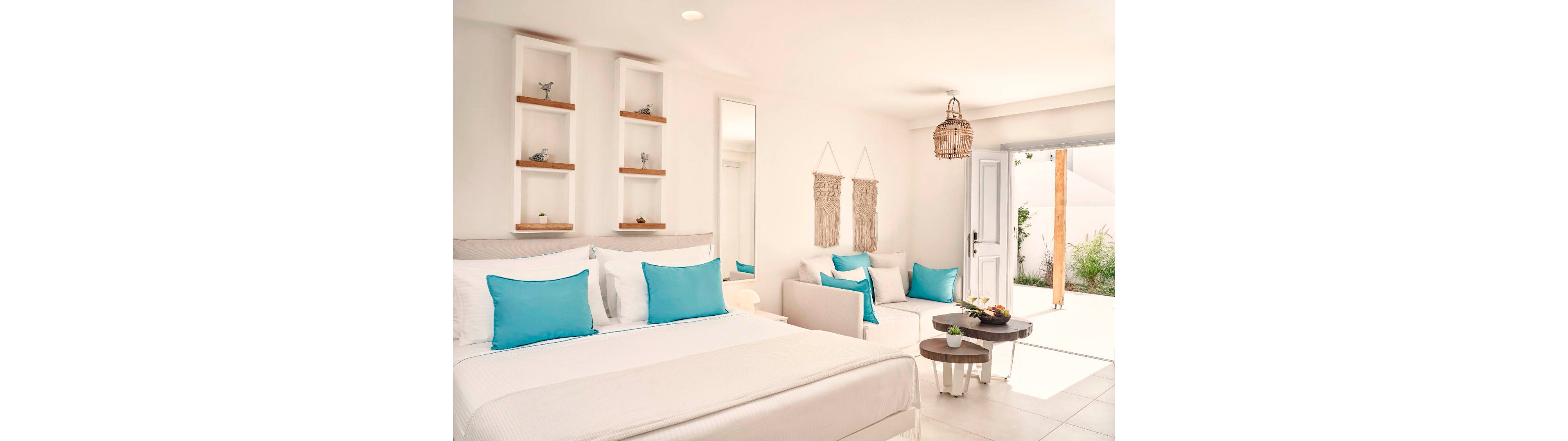 A modern, airy bedroom features a large bed with crisp white linens and turquoise pillows. A small seating area with a sofa and matching cushions faces a set of round nesting tables holding decorative items. Built-in wall shelves, woven wall hangings, and a rattan pendant light add natural texture to the minimalist decor. Sunlight streams in through an open door, revealing greenery outside and enhancing the fresh, coastal atmosphere.